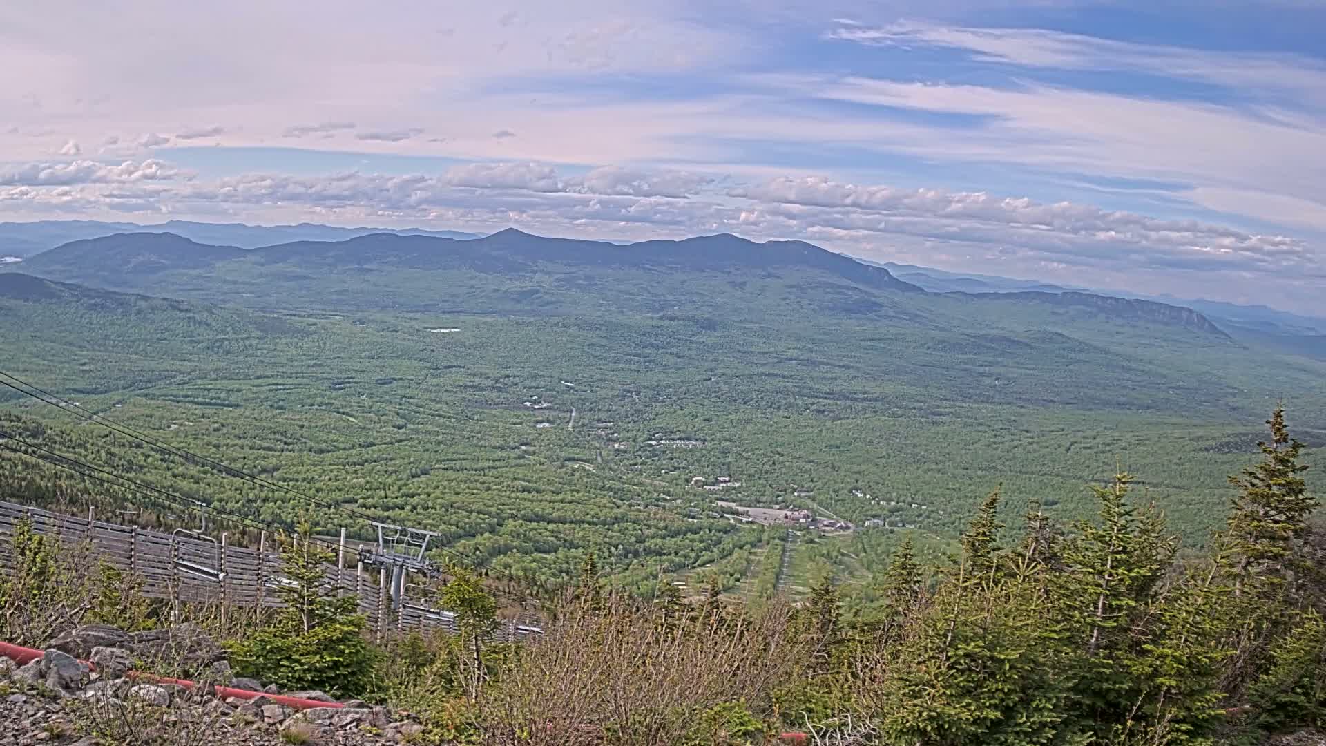 A mountaintop view on a partly cloudy day shows a vast expanse of green forest stretching to distant, hazy blue mountains, with a ski lift and snowmaking system partially visible in the foreground.