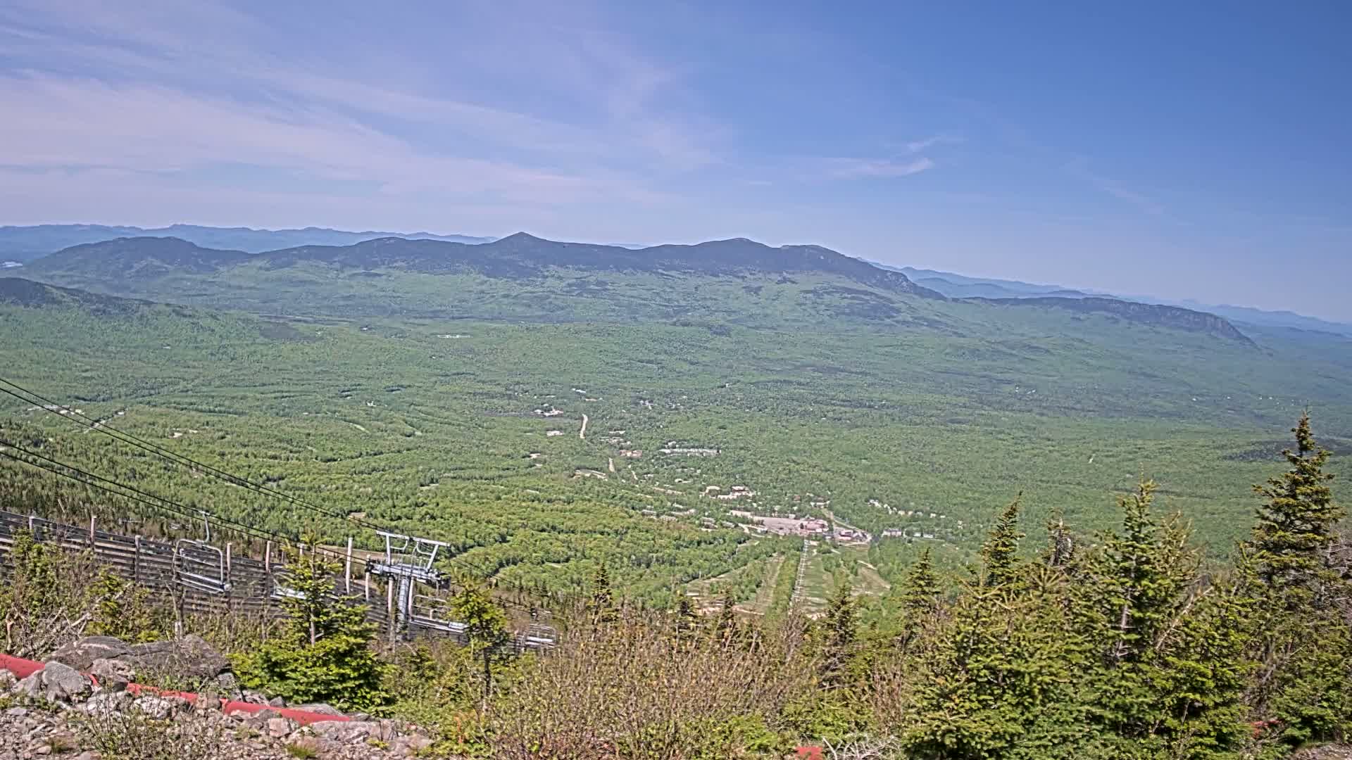 A high-angle, long shot on a clear, sunny day shows a vast expanse of green, forested mountains and valleys, with a ski lift visible in the foreground.