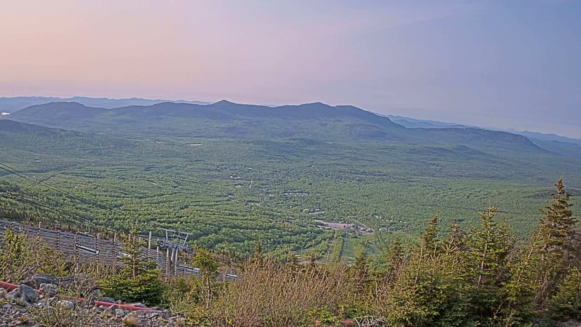 A vast expanse of lush green forest and distant mountains is viewed from a high vantage point under a clear, pale sky.