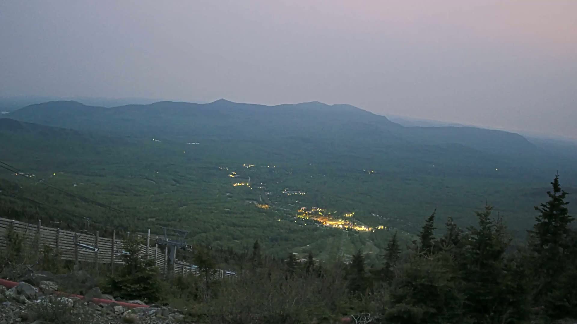 A hazy, twilight view from a mountaintop shows a valley of green forests with a small town illuminated below, and a ski lift partially visible in the foreground.