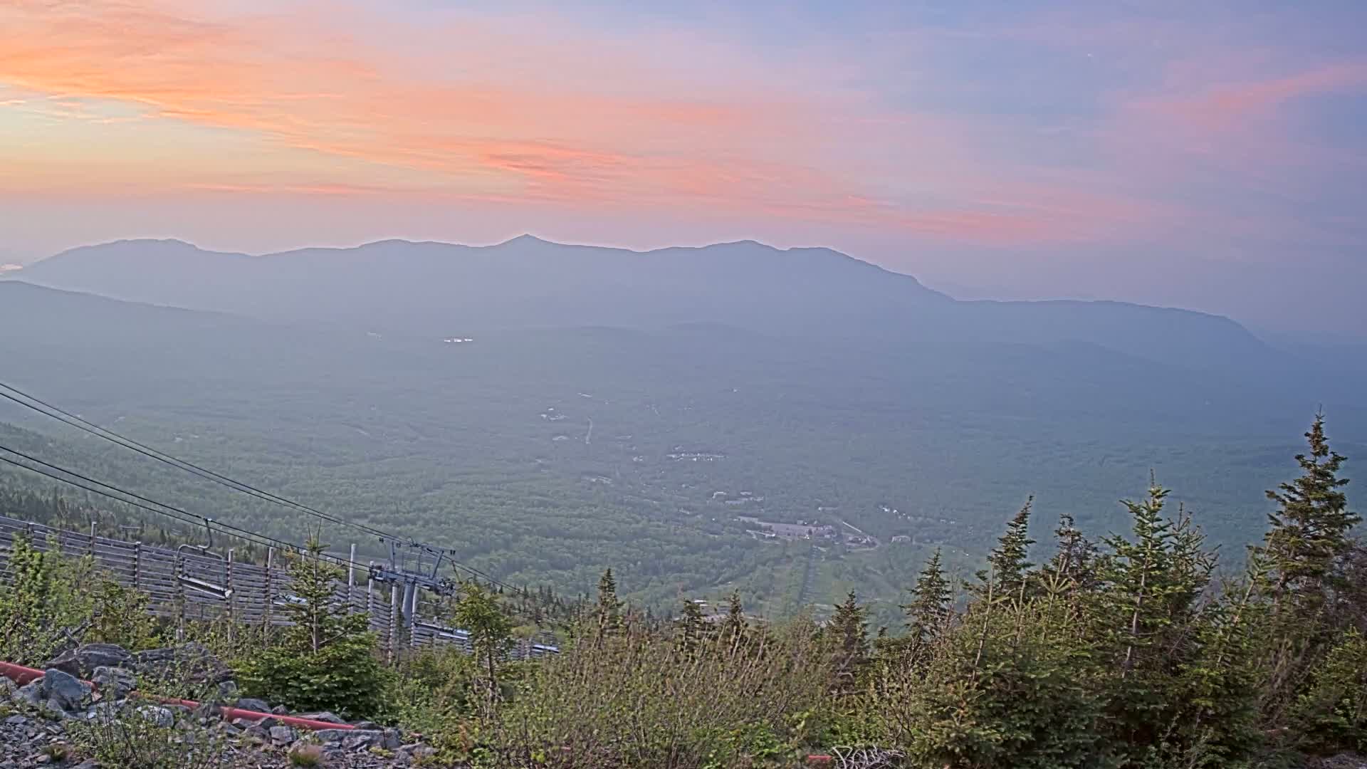 A hazy, pastel sunrise illuminates a mountainous landscape, viewed from a high vantage point with a ski lift in the foreground.