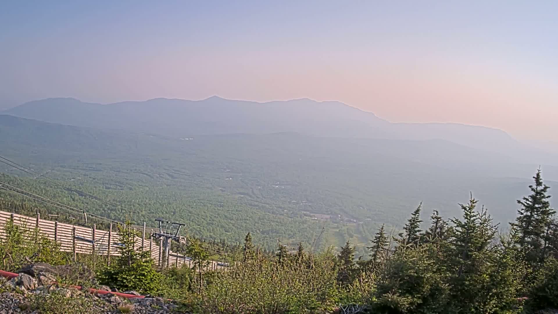 A hazy, mountainous landscape is visible from a high vantage point, showing a chairlift and wooden fence in the foreground.