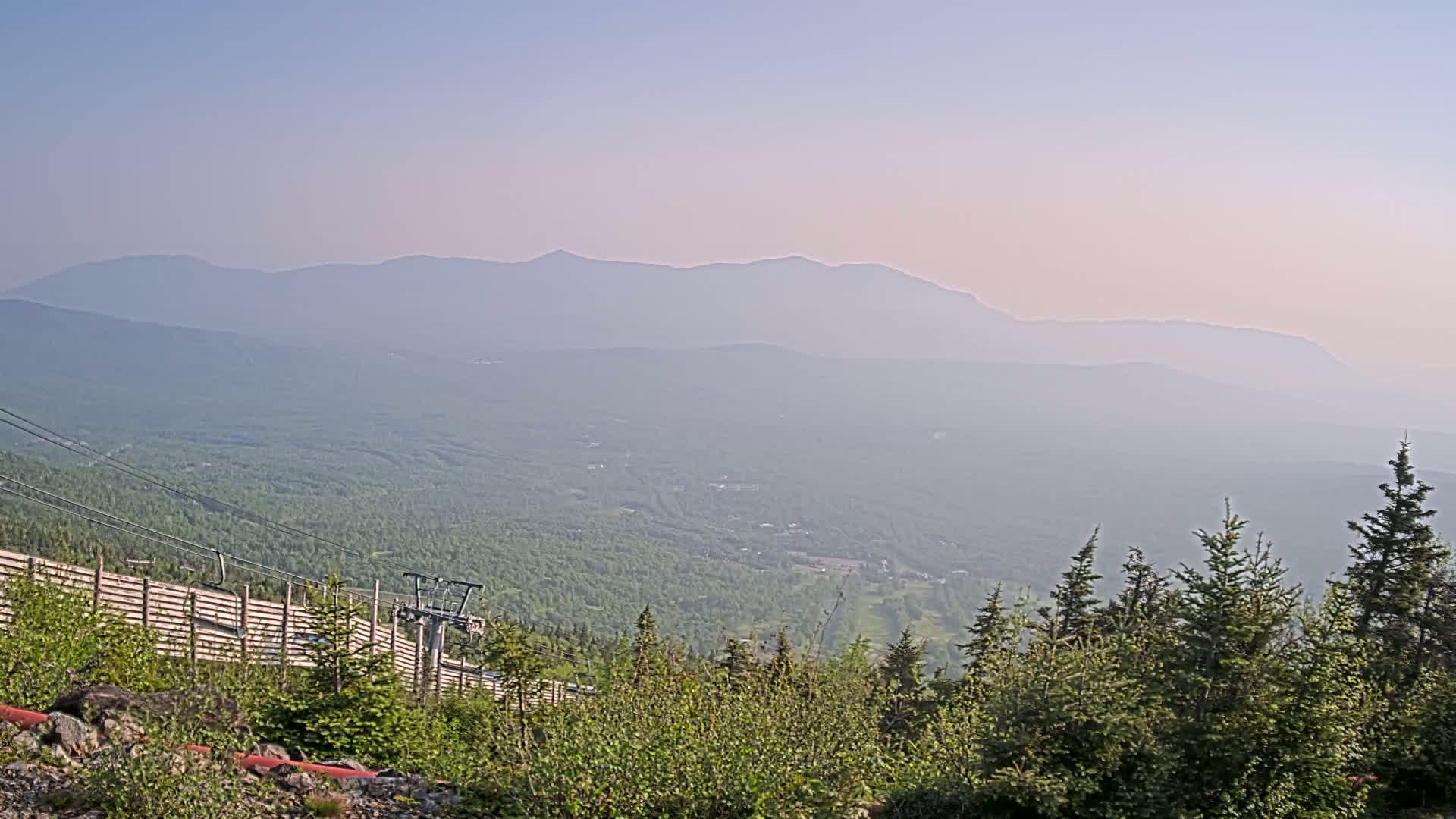 A hazy, mountainous landscape is viewed from a high vantage point, showing a chairlift and a wooden fence near evergreen trees in the foreground.