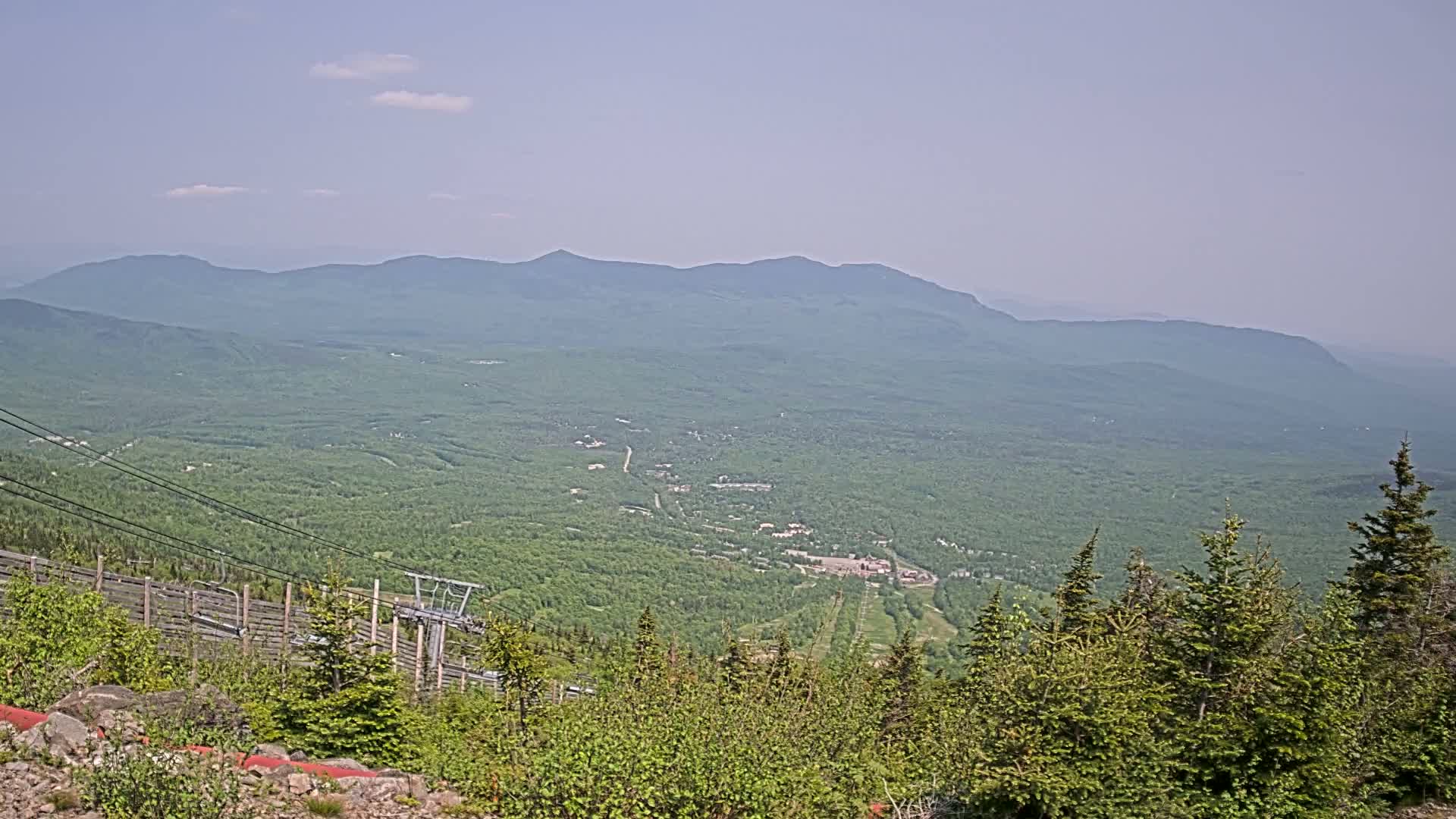 A hazy, sunny day reveals a vast, verdant mountain valley viewed from a high vantage point, featuring a chairlift and a distant mountain range.
