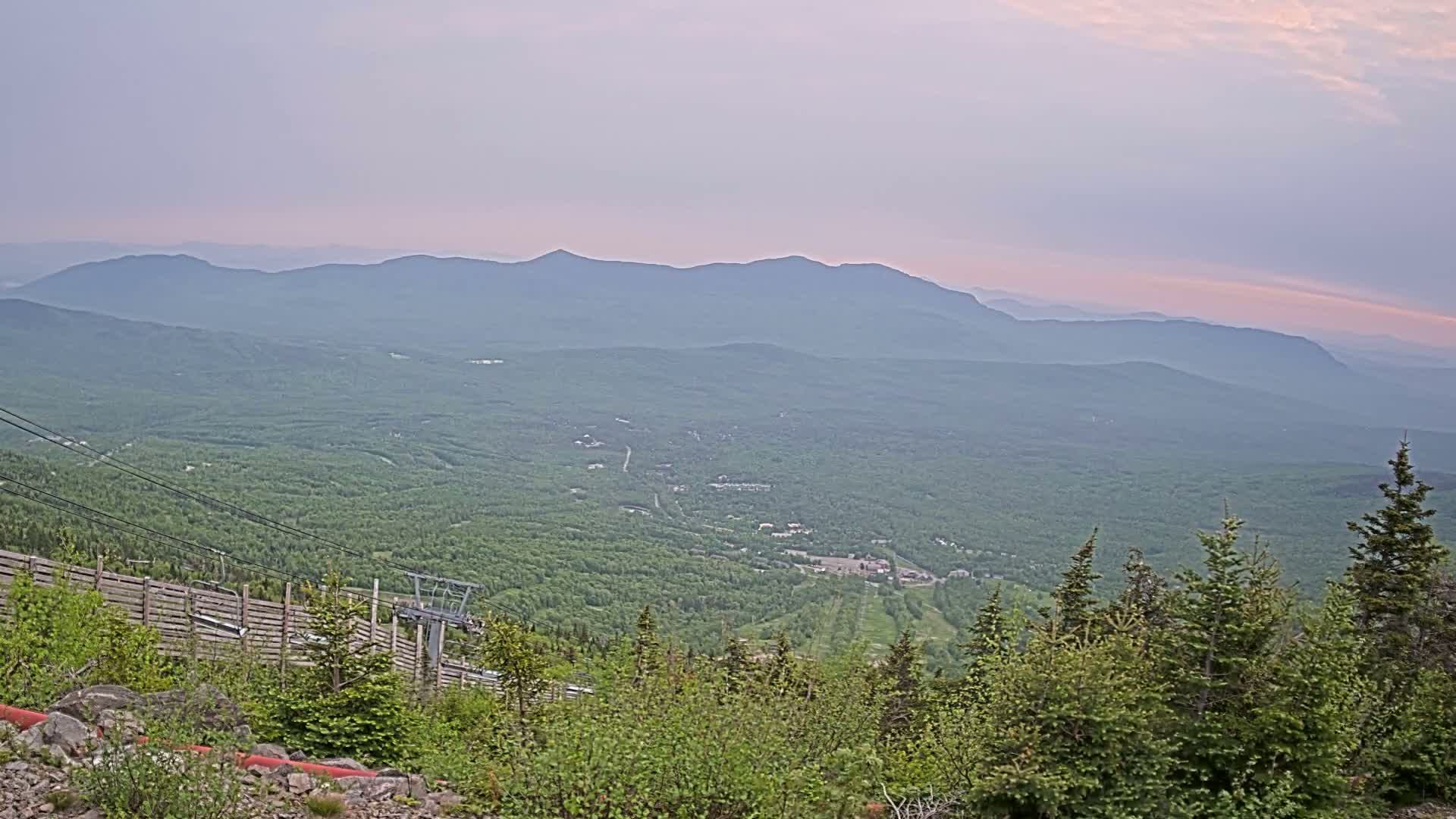 A hazy, sunset view from a mountaintop shows a vast expanse of green forest and distant, layered mountains.