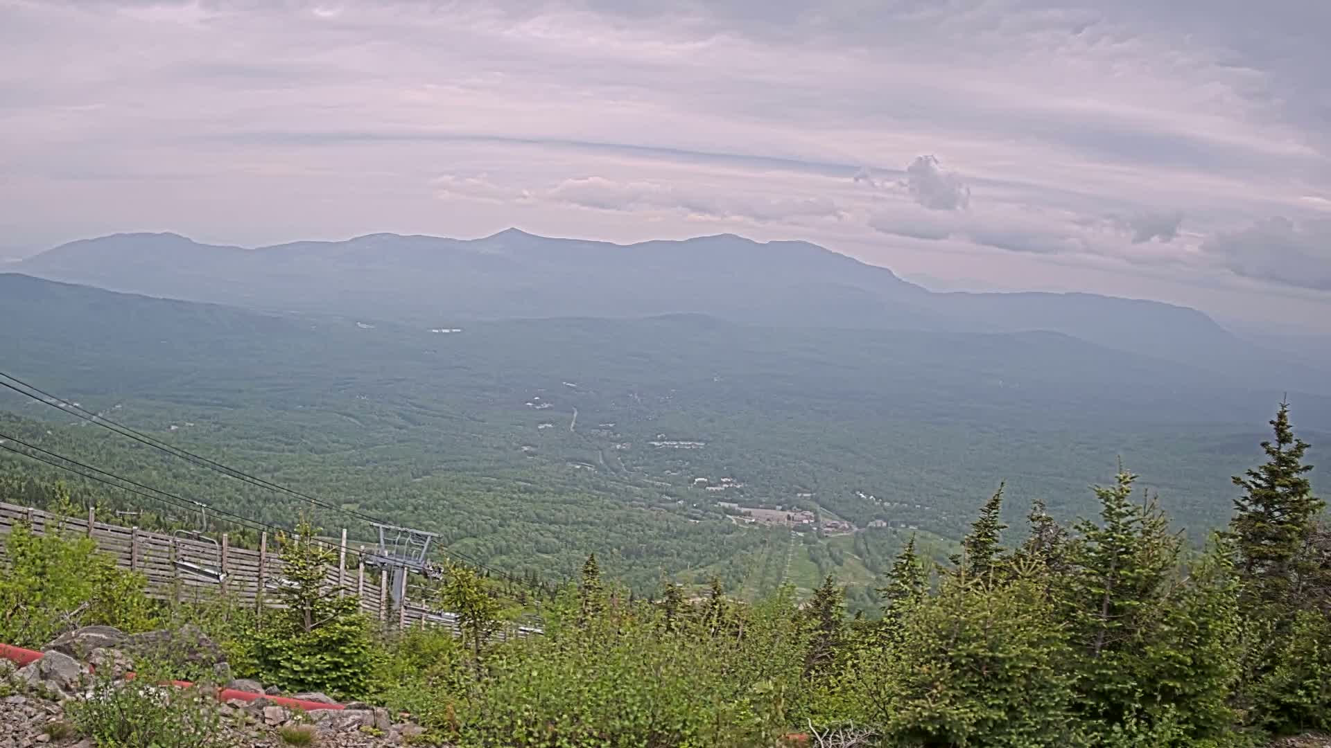 A mountaintop view on an overcast day shows a chairlift, a vast expanse of green forest, and distant, hazy mountains.