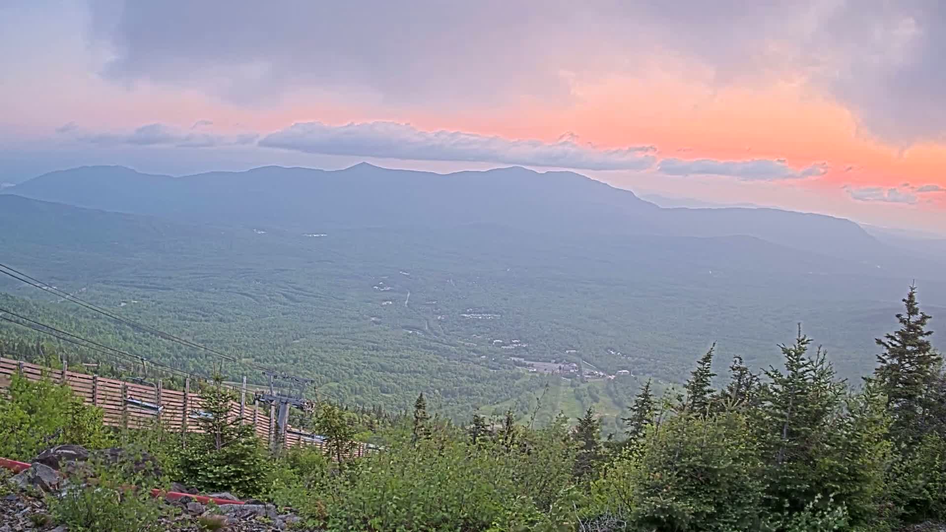 A mountaintop view at sunset shows a vast expanse of green forests and distant mountains under a pale pink and purple sky.