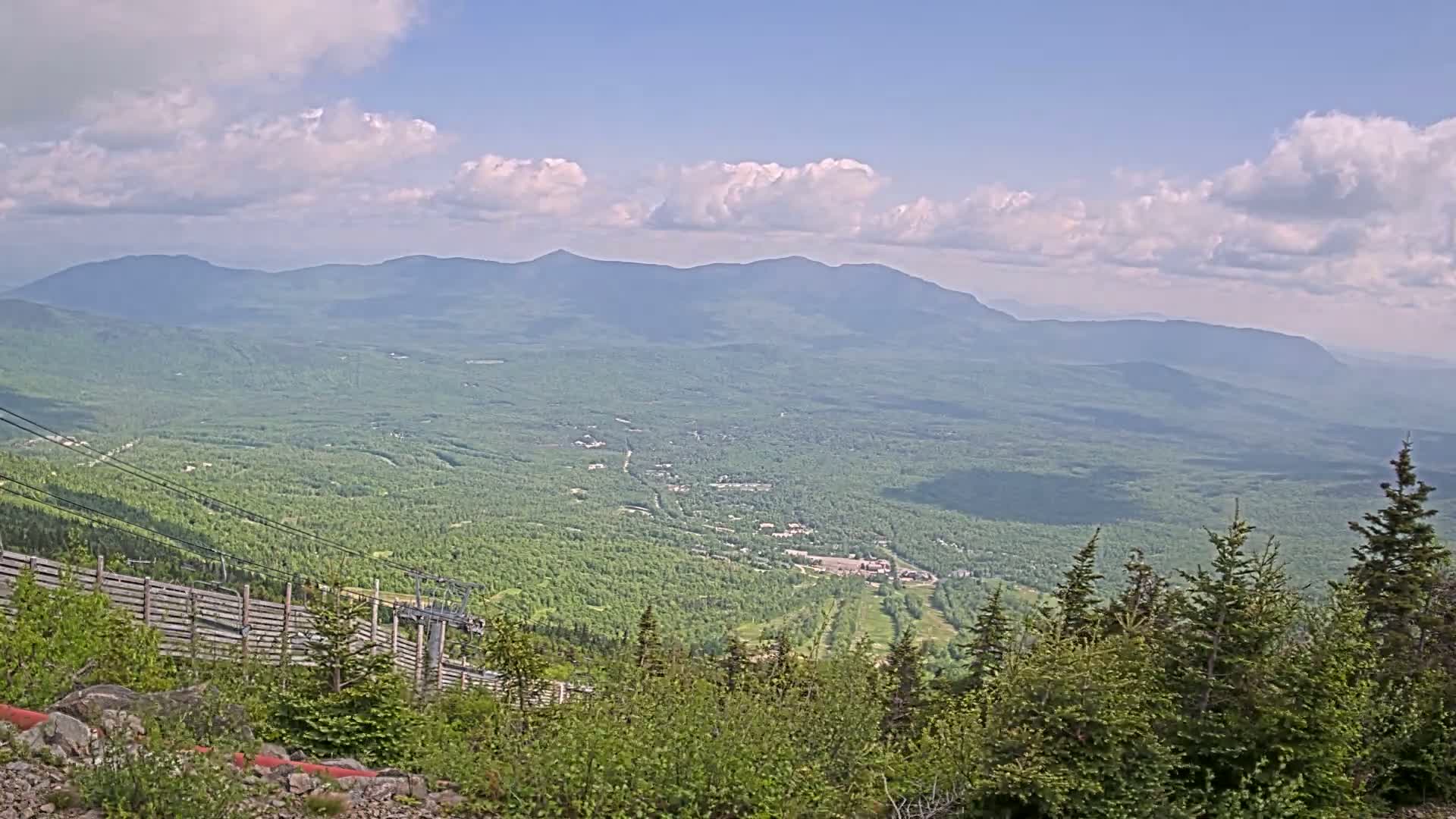 A partly cloudy day reveals a vast, verdant valley stretching to distant mountains, with a ski lift and wooded slopes in the foreground.