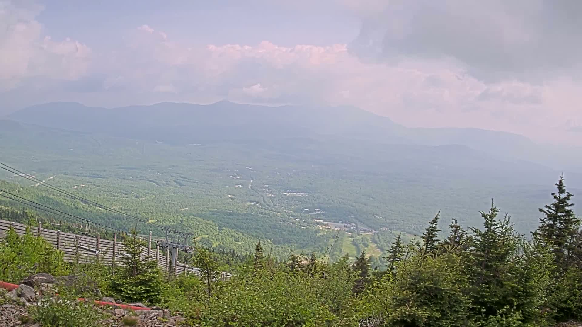 A hazy, partly cloudy day reveals a vast expanse of green mountains and forests, with a ski lift visible in the foreground.