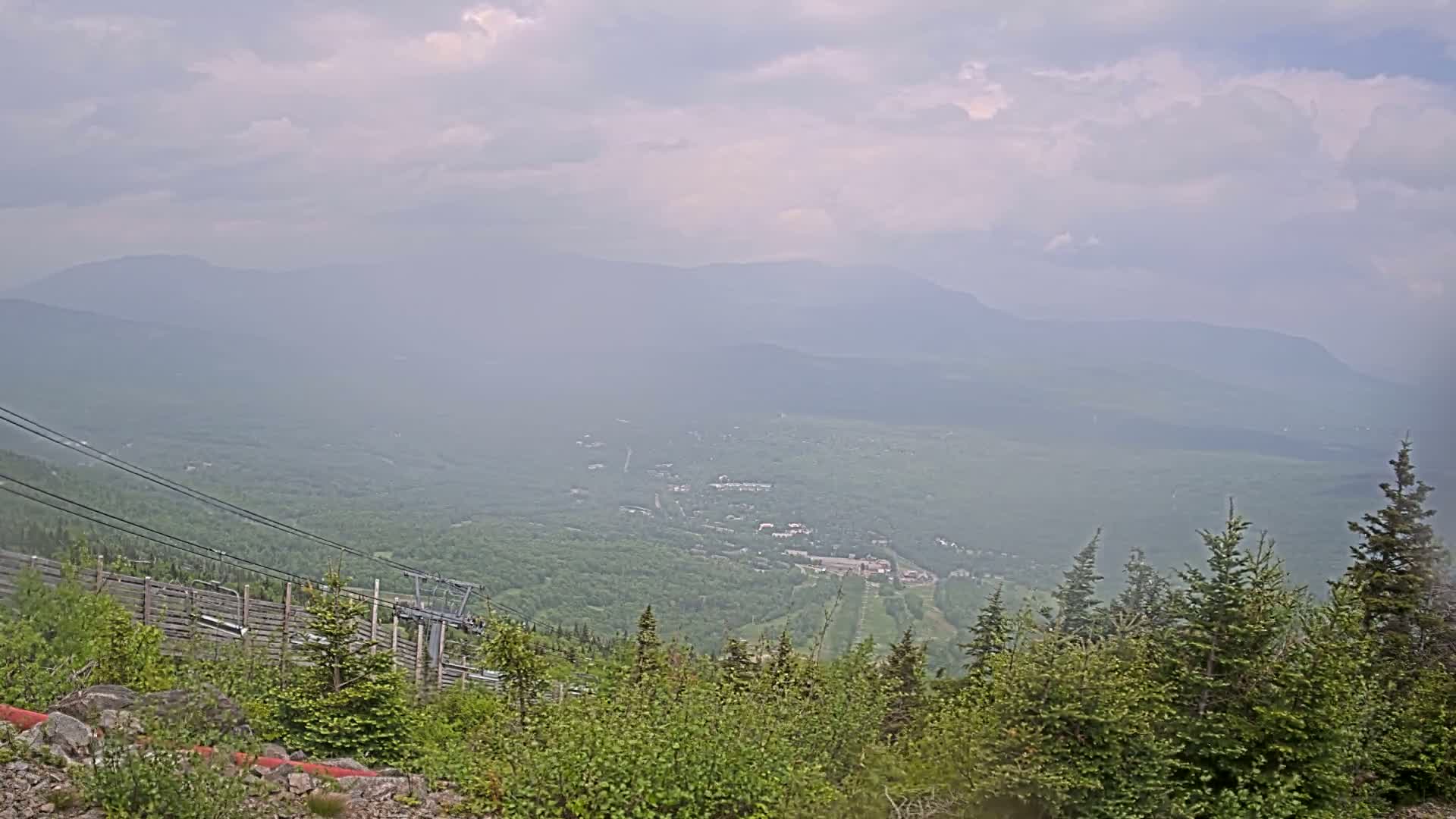 A hazy mountain valley view shows a ski lift and a forested hillside in the foreground, with distant mountains under a cloudy sky.