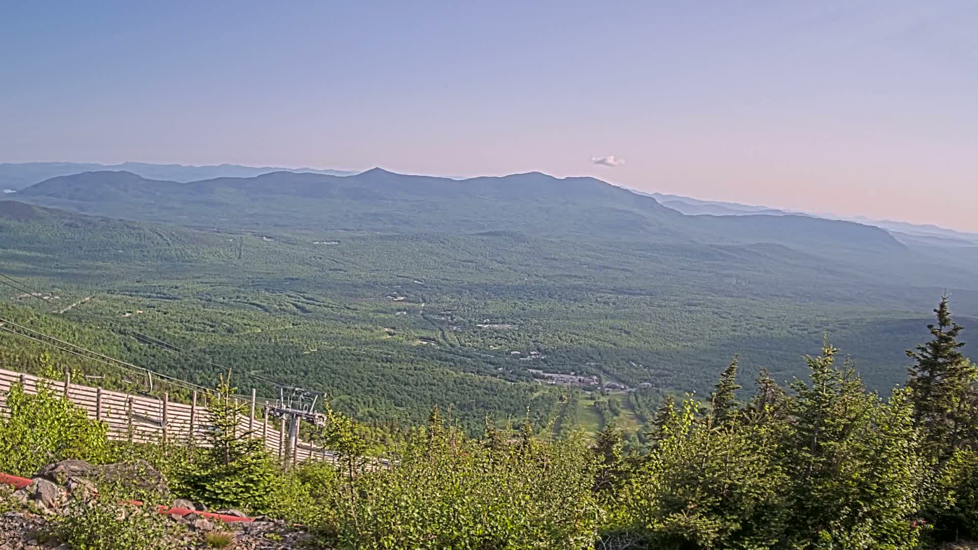 A high-angle, long shot of a vast, forested mountain range under a clear, light blue sky shows a chairlift and ski area in the foreground.
