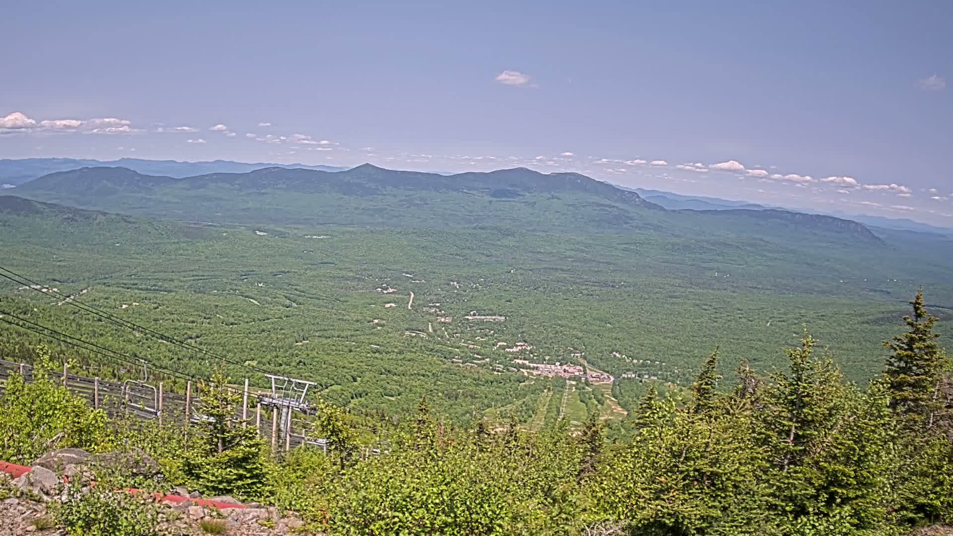 A mountaintop view on a partly sunny day reveals a vast expanse of green forest stretching to distant, hazy mountains, with a ski lift and chair visible in the foreground.