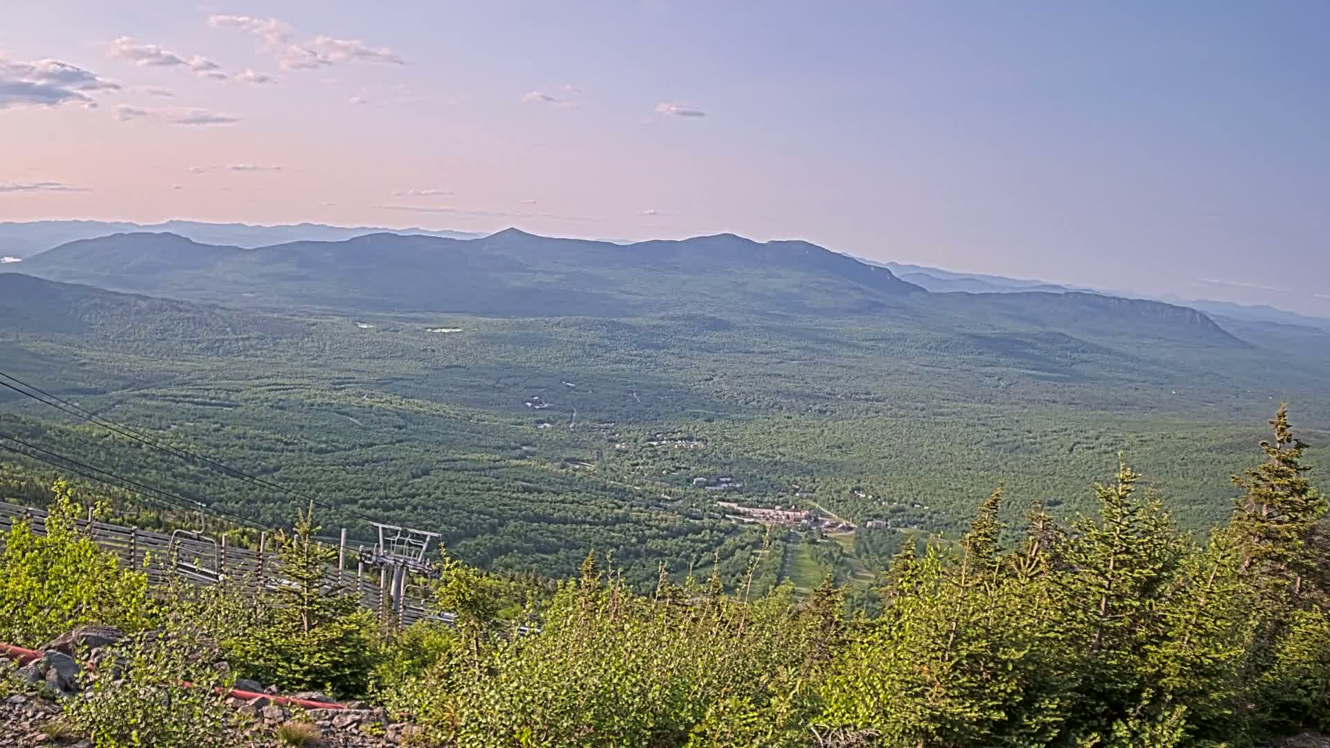 A mountaintop view on a partly sunny day reveals a vast expanse of green forest stretching to distant, hazy mountains, with a ski lift and chair visible in the foreground.