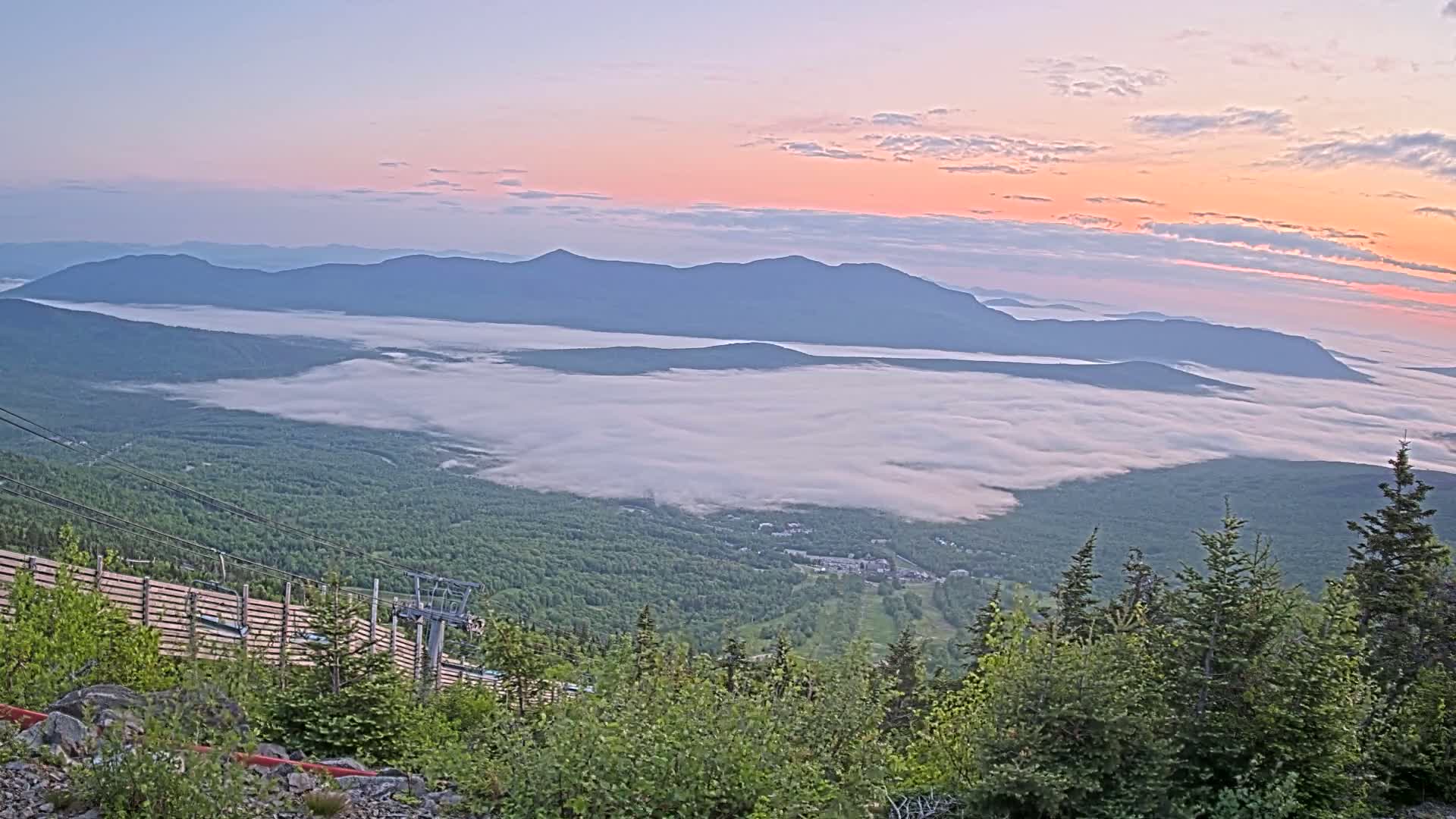 A sunrise casts a pink and orange glow over a valley shrouded in fog, with mountains in the background and a ski lift visible in the foreground.