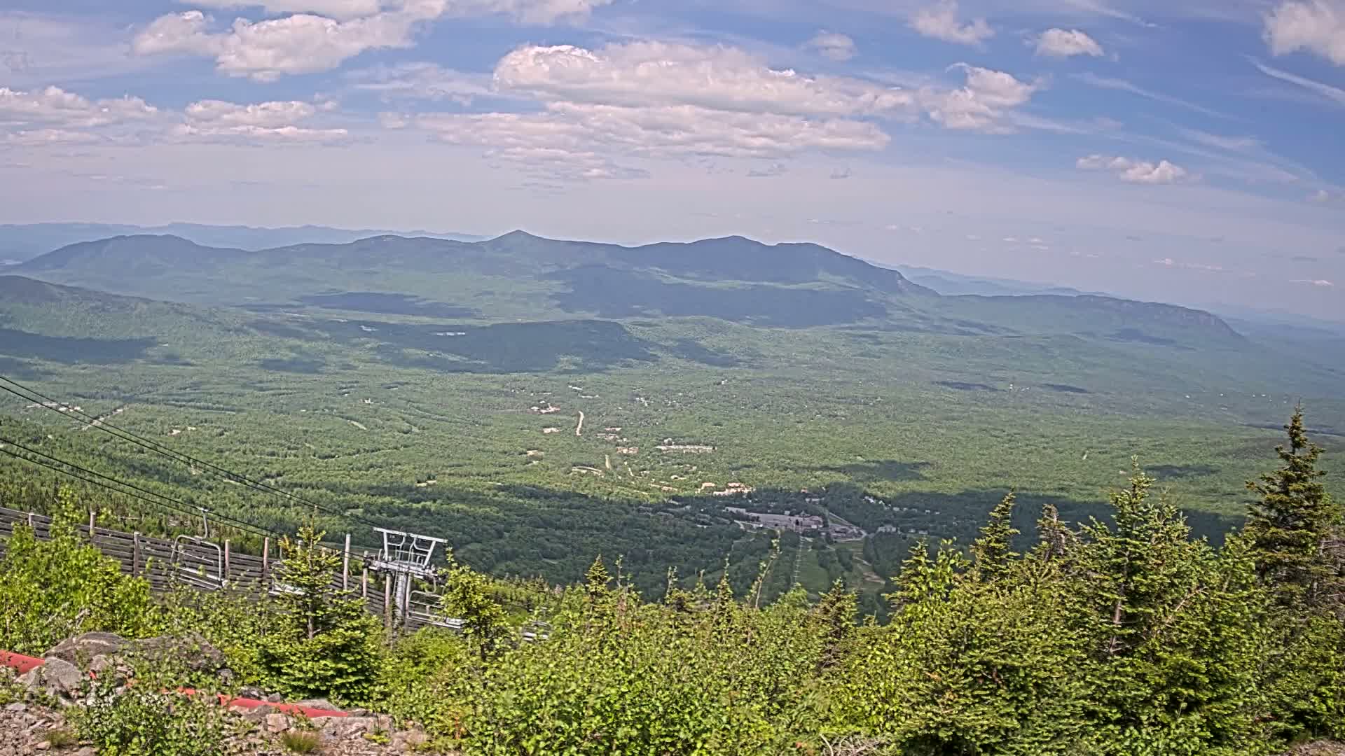 A vast expanse of lush green mountains and valleys stretches to the horizon under a partly cloudy blue sky, with a ski lift partially visible in the foreground.