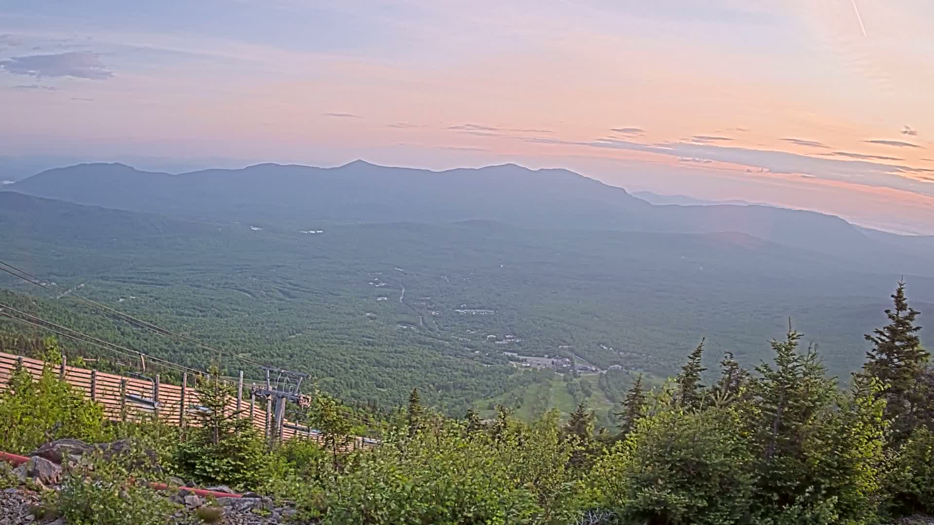A mountaintop view at sunset shows a vast expanse of green forest stretching to distant, hazy mountains under a pale pink and lavender sky.