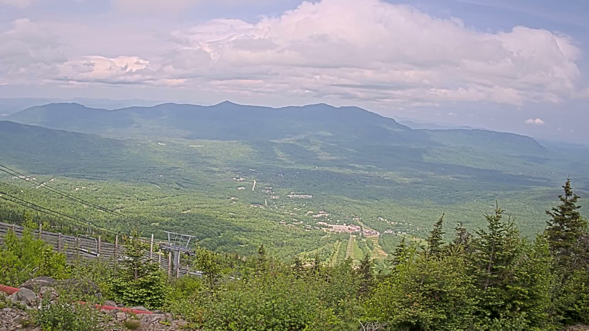 A mostly cloudy day reveals a vast, green mountain valley with a ski resort visible in the distance, seen from a high vantage point near a chairlift.