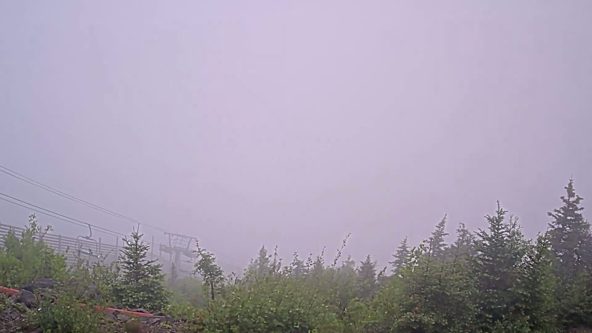 A foggy scene shows a ski lift partially obscured by thick fog and low-lying vegetation.