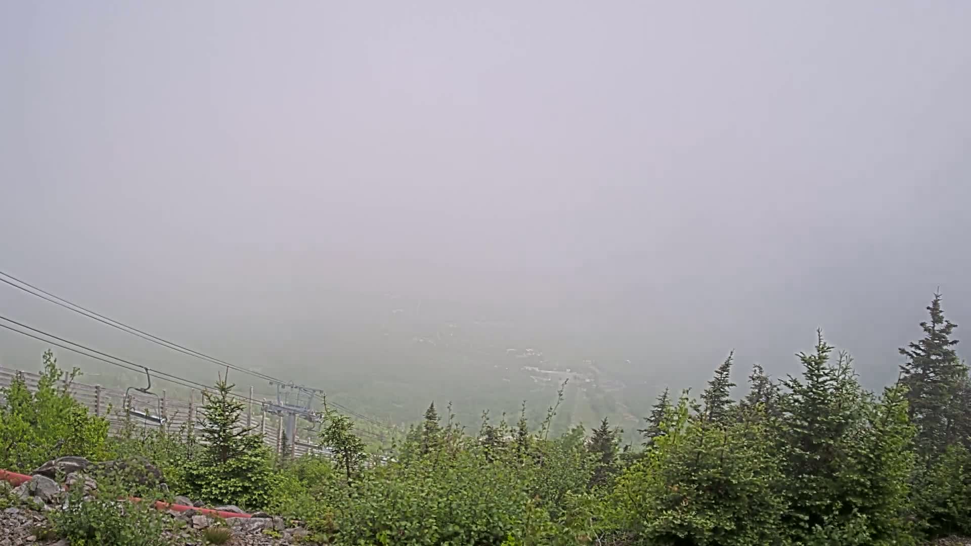 A foggy mountain vista is partially obscured by a thick fog, showing a chairlift and evergreen trees in the foreground.