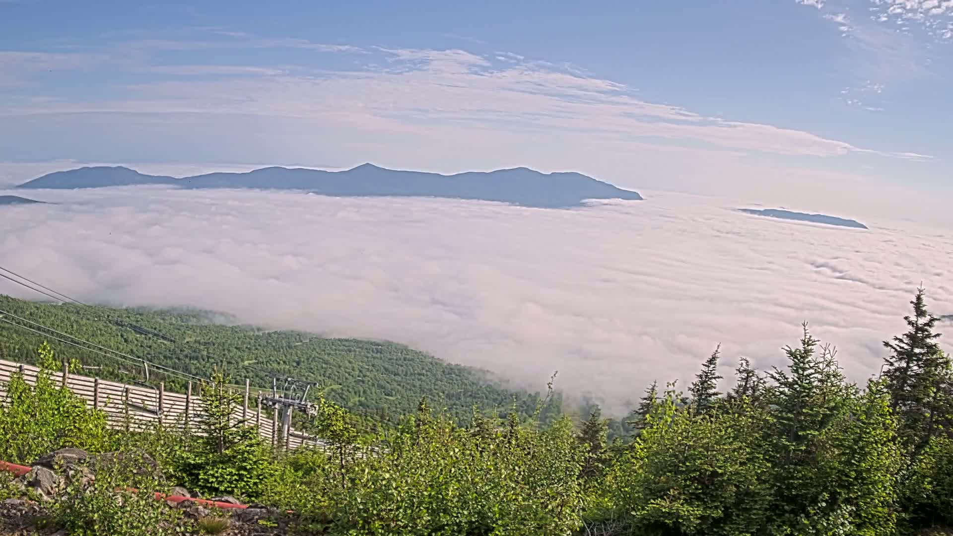 A mountaintop view on a clear day shows a vast expanse of low-lying clouds covering a valley and distant mountains.
