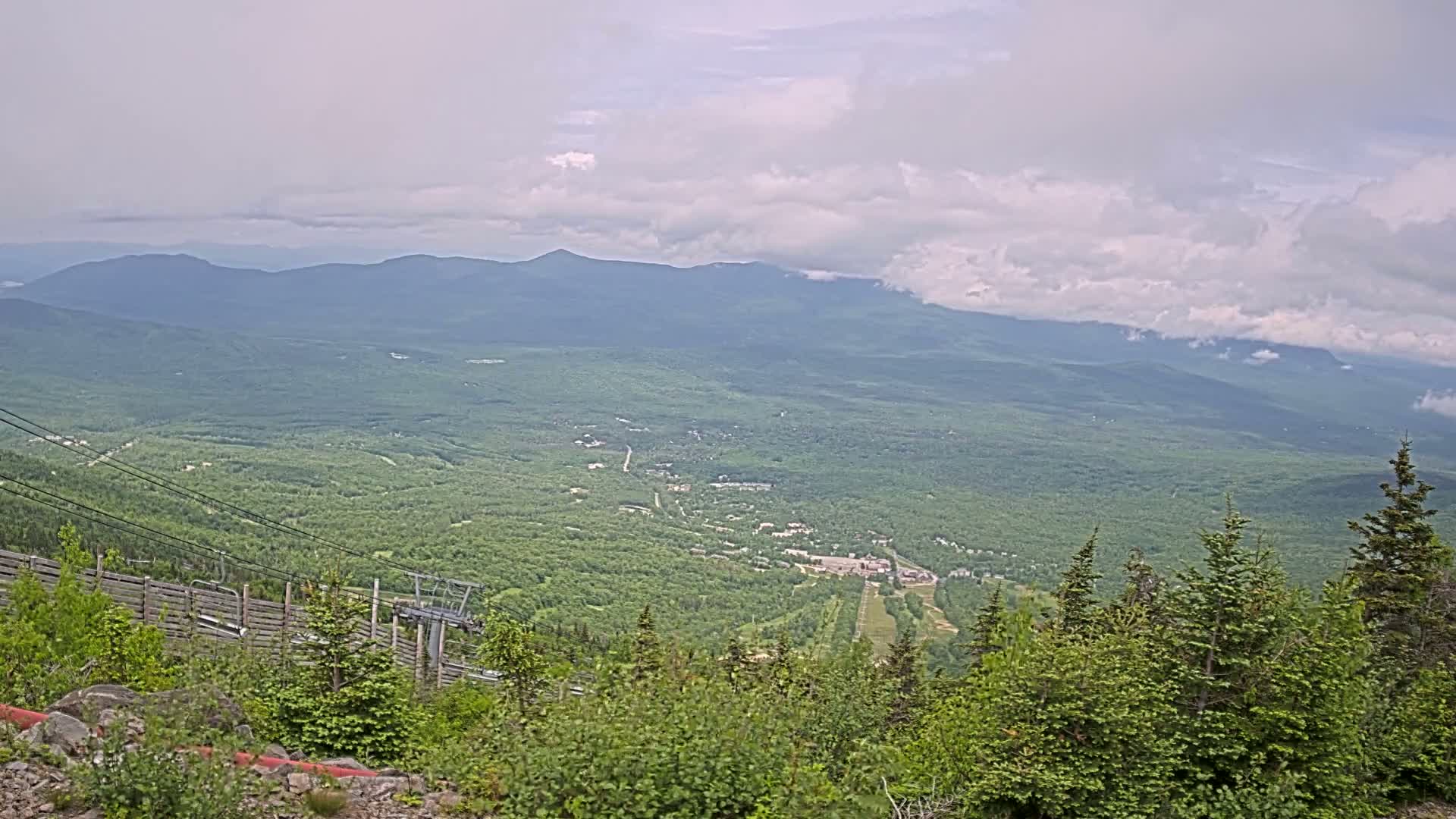 A high-angle view of a vast, green mountain valley under a cloudy sky, showing a ski resort in the mid-ground and a ski lift in the foreground.