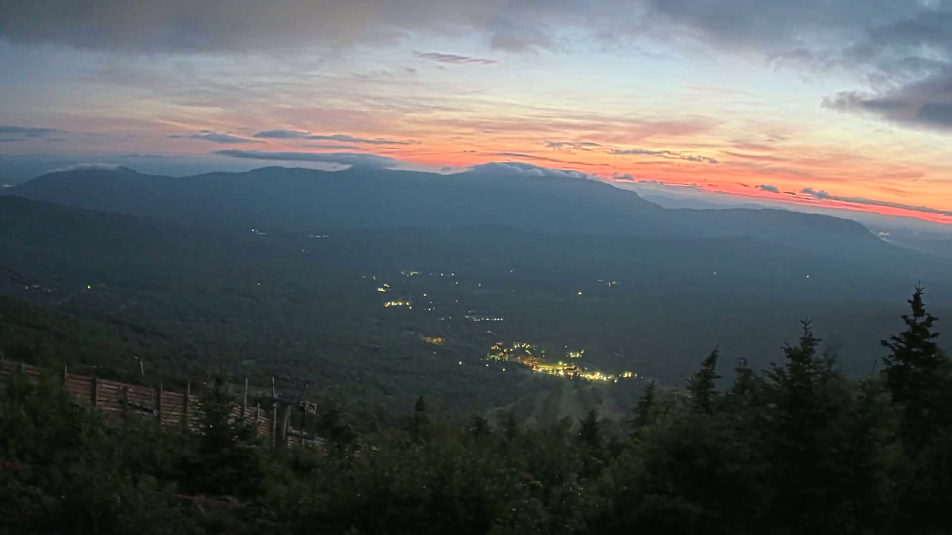 A mountain vista at sunset shows a valley with scattered lights below, under a partly cloudy, pinkish sky.