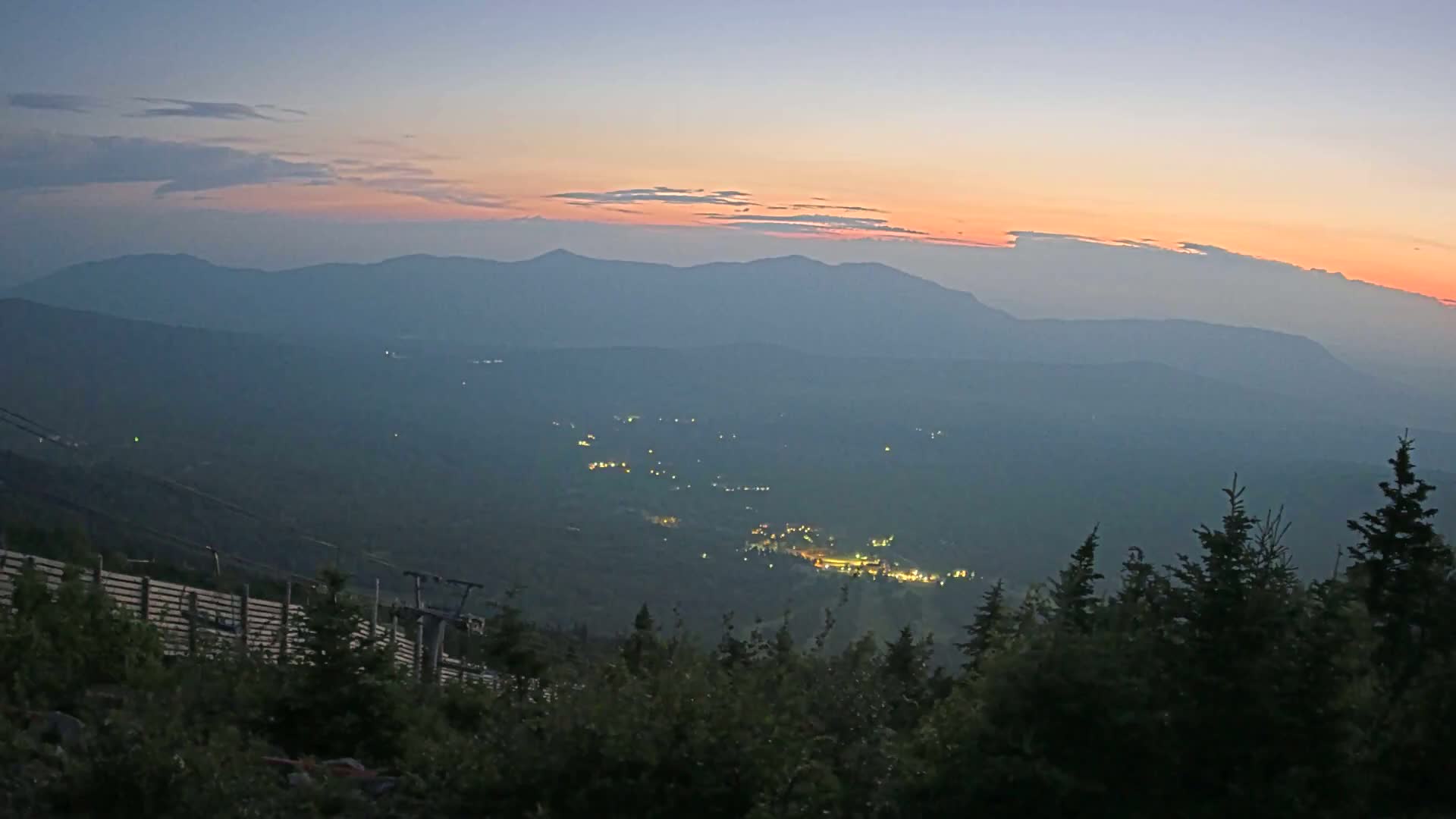 A hazy sunset illuminates a valley with scattered lights below a mountain range, viewed from a high vantage point with evergreen trees in the foreground.