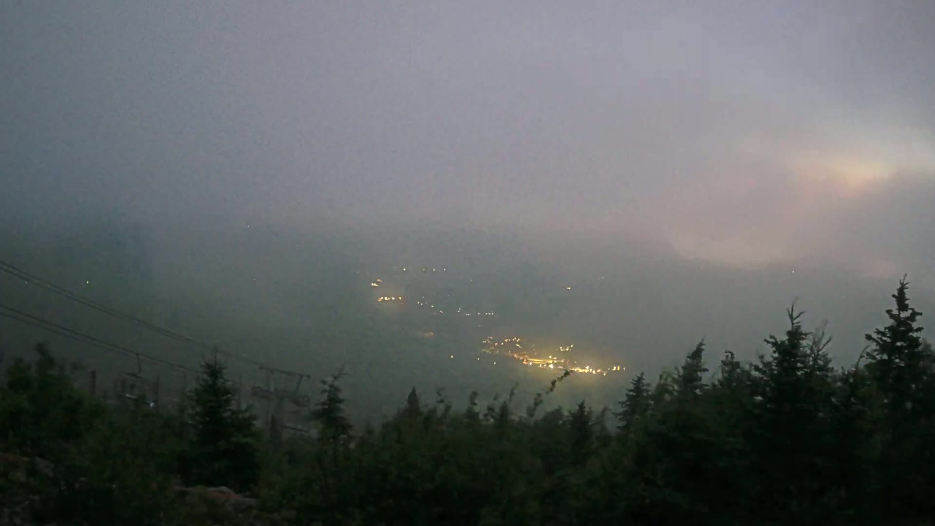 Faint lights from a distant settlement are barely visible through a thick fog hanging over a dark, forested hillside.