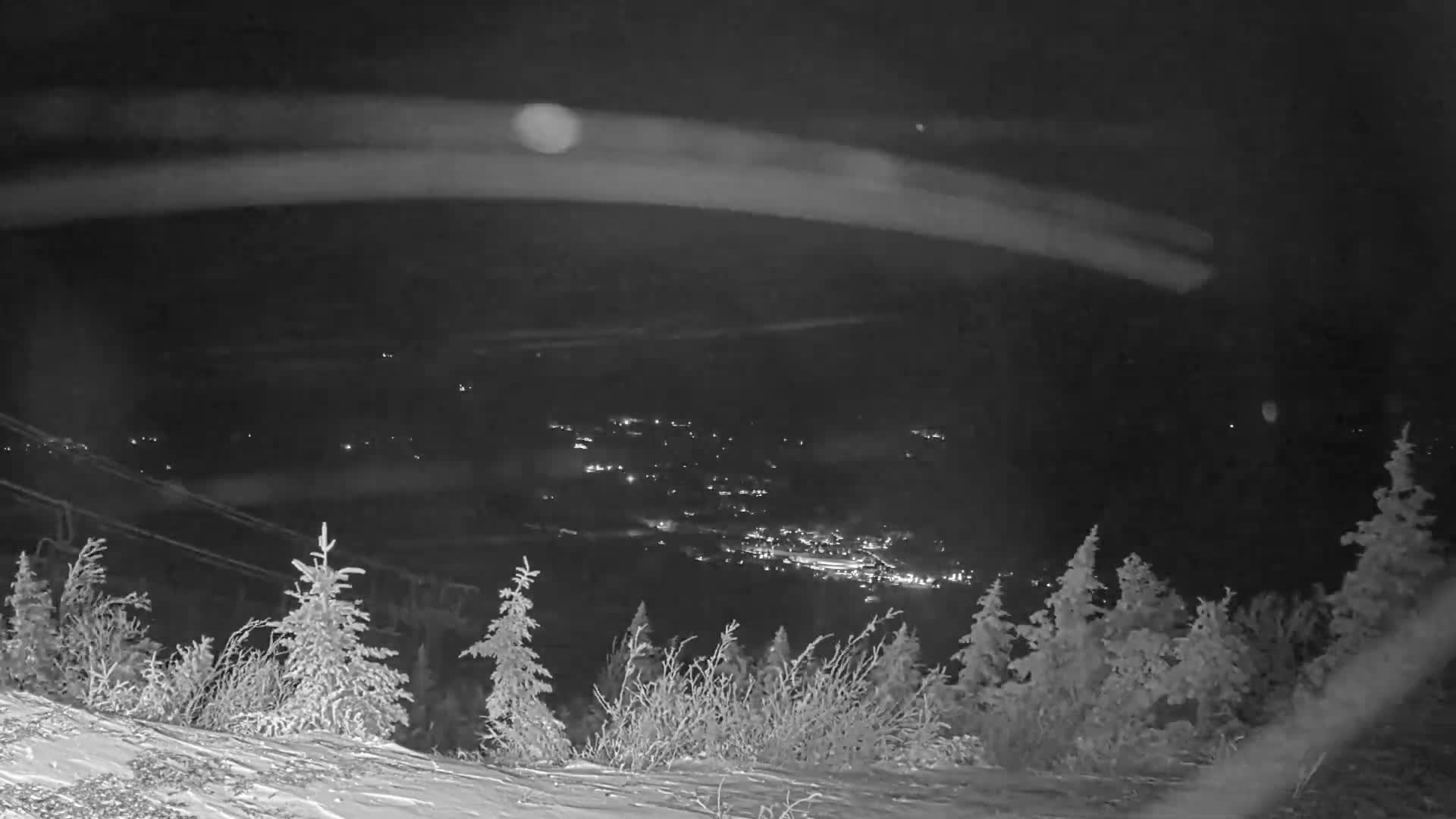 A black and white night shot from a snow-covered slope reveals a distant town twinkling with lights in a valley, under clear and cold winter conditions.