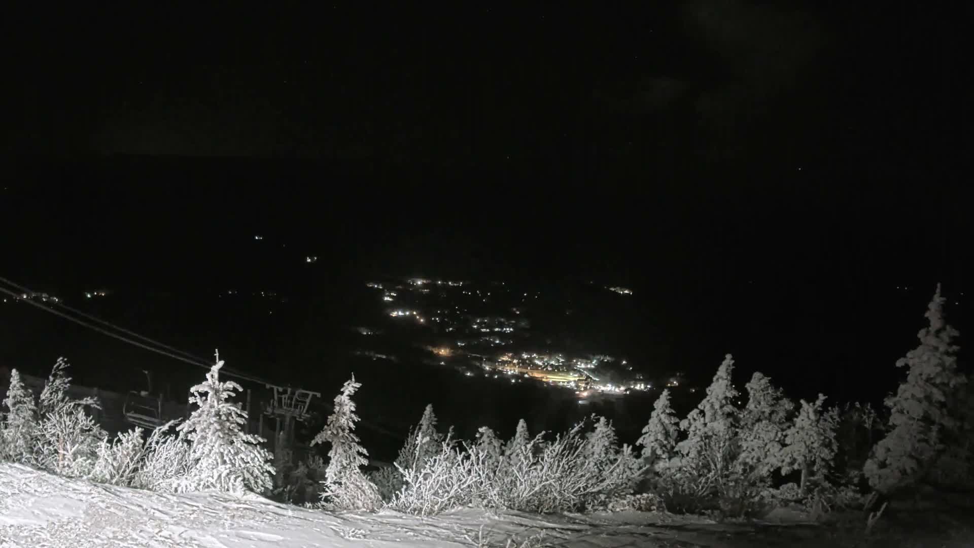 A clear, cold winter night scene on a snowy mountain slope features frost-laden trees and a ski lift in the foreground, overlooking a distant valley town twinkling with lights.