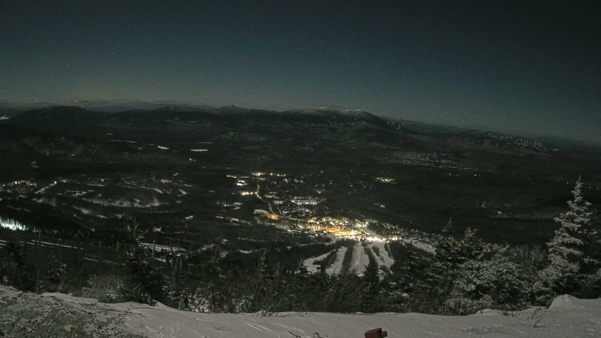A clear, snowy night offers a panoramic view from a mountain, revealing a brightly lit town and ski slopes nestled among dark forests and distant peaks.