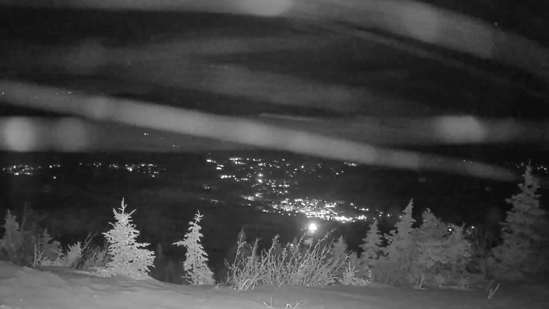 A clear, snowy night offers a panoramic view from a mountain, revealing a brightly lit town and ski slopes nestled among dark forests and distant peaks.