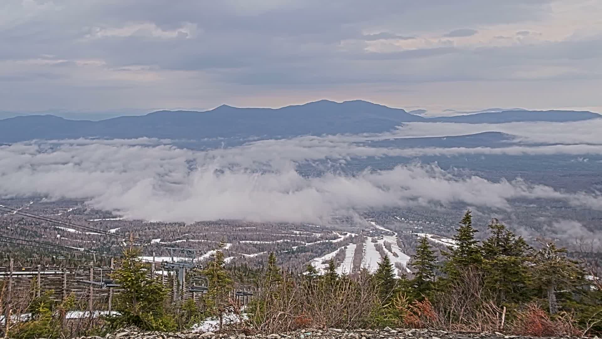A clear, snowy night offers a panoramic view from a mountain, revealing a brightly lit town and ski slopes nestled among dark forests and distant peaks.