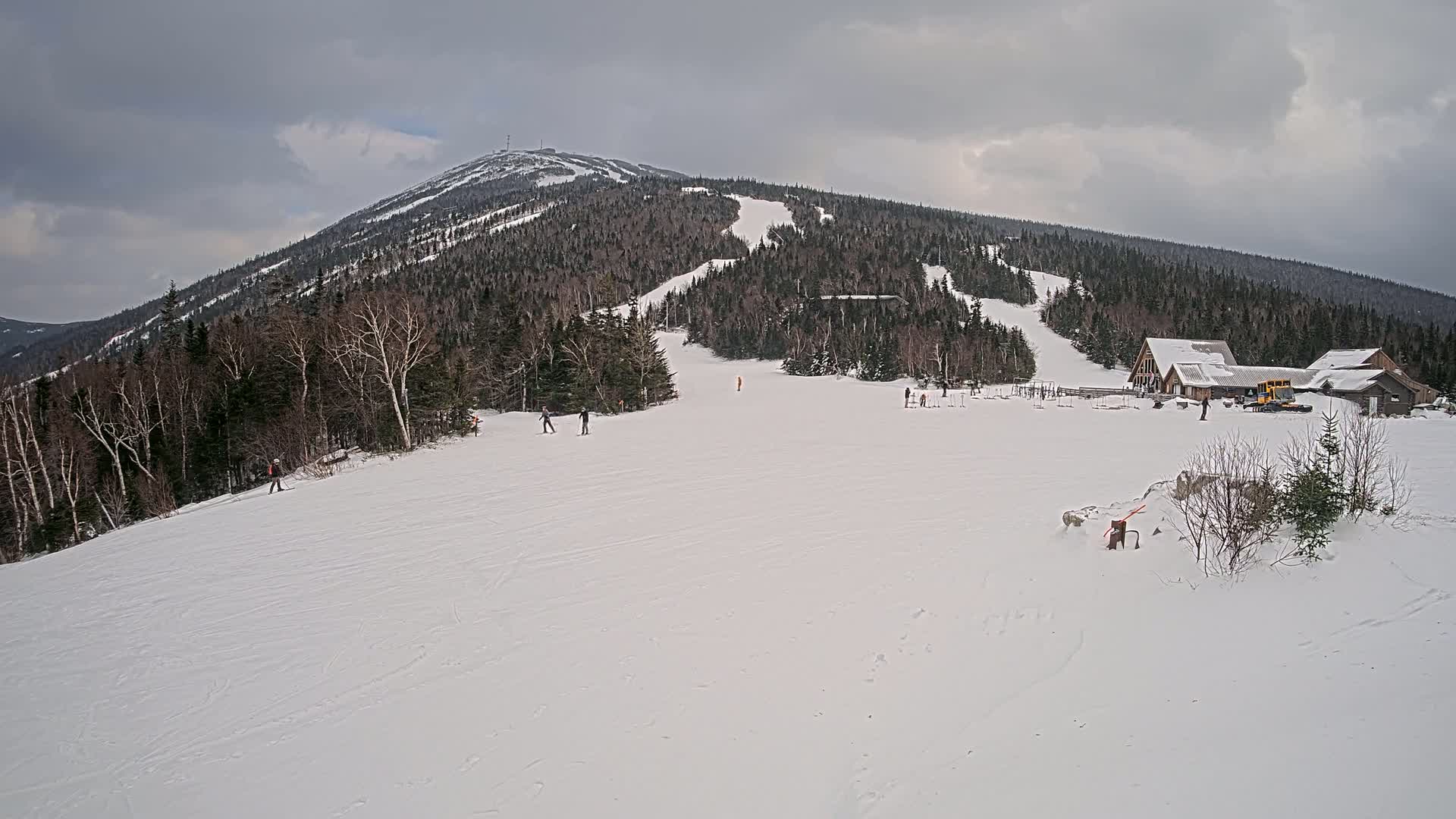 A snow-covered mountain slope with several skiers, a base lodge, and a snowcat under an overcast sky.