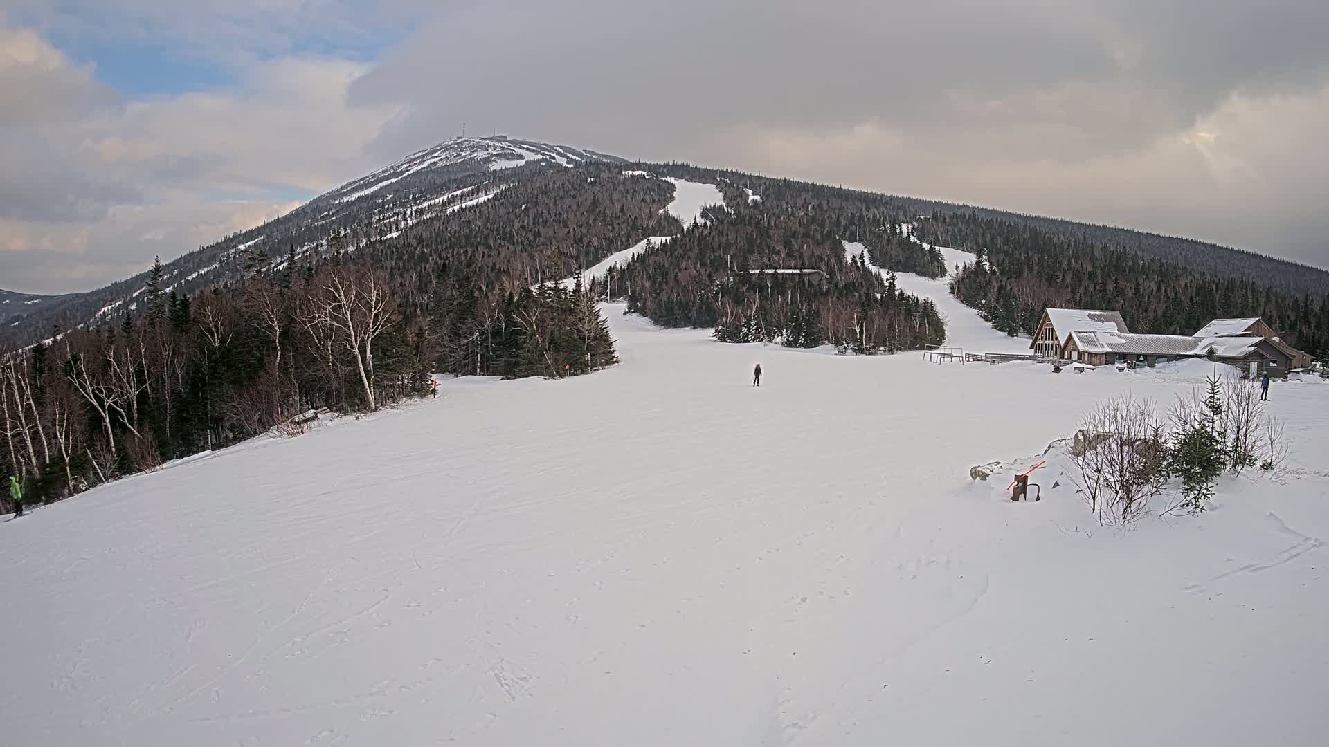 A snow-covered ski slope stretches out before a large mountain, under a cloudy sky.