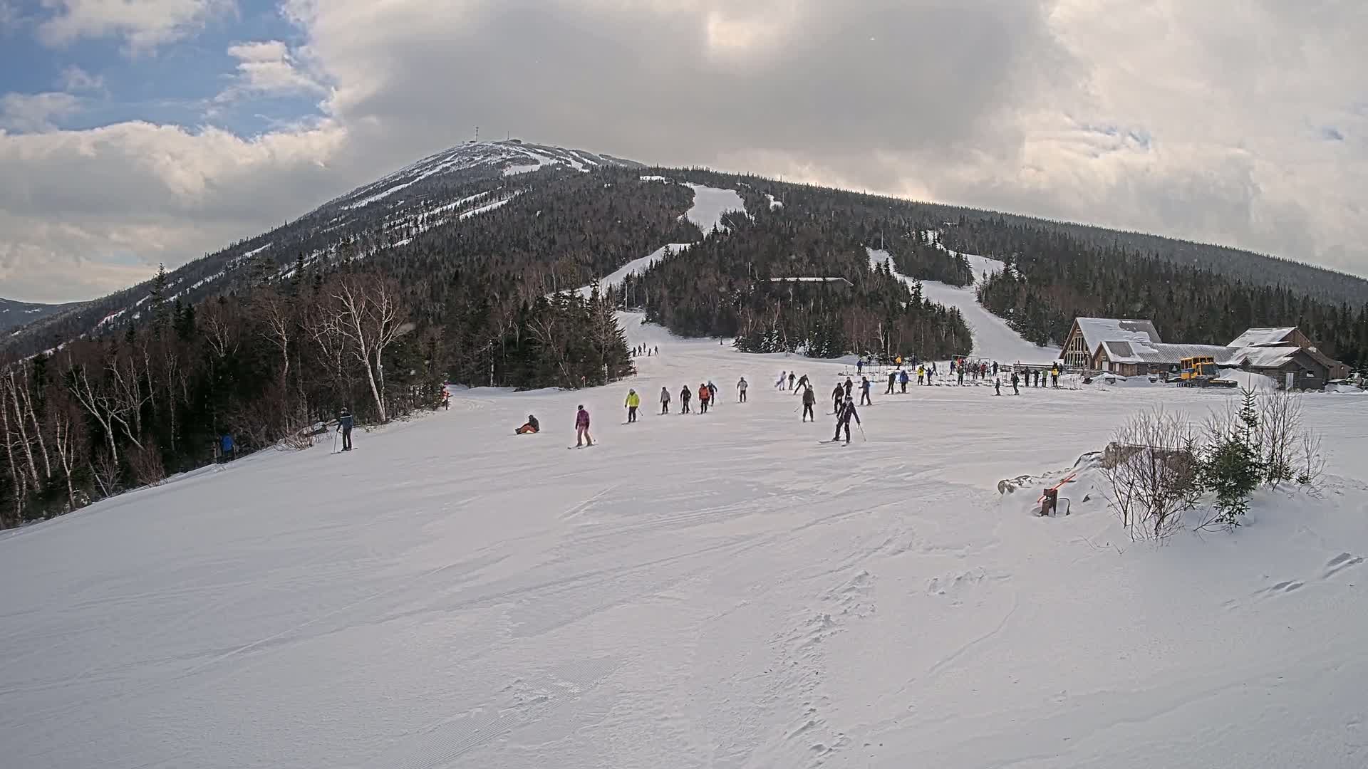A snowy ski slope with several skiers and snowboarders is situated below a snow-covered mountain under a partly cloudy sky.