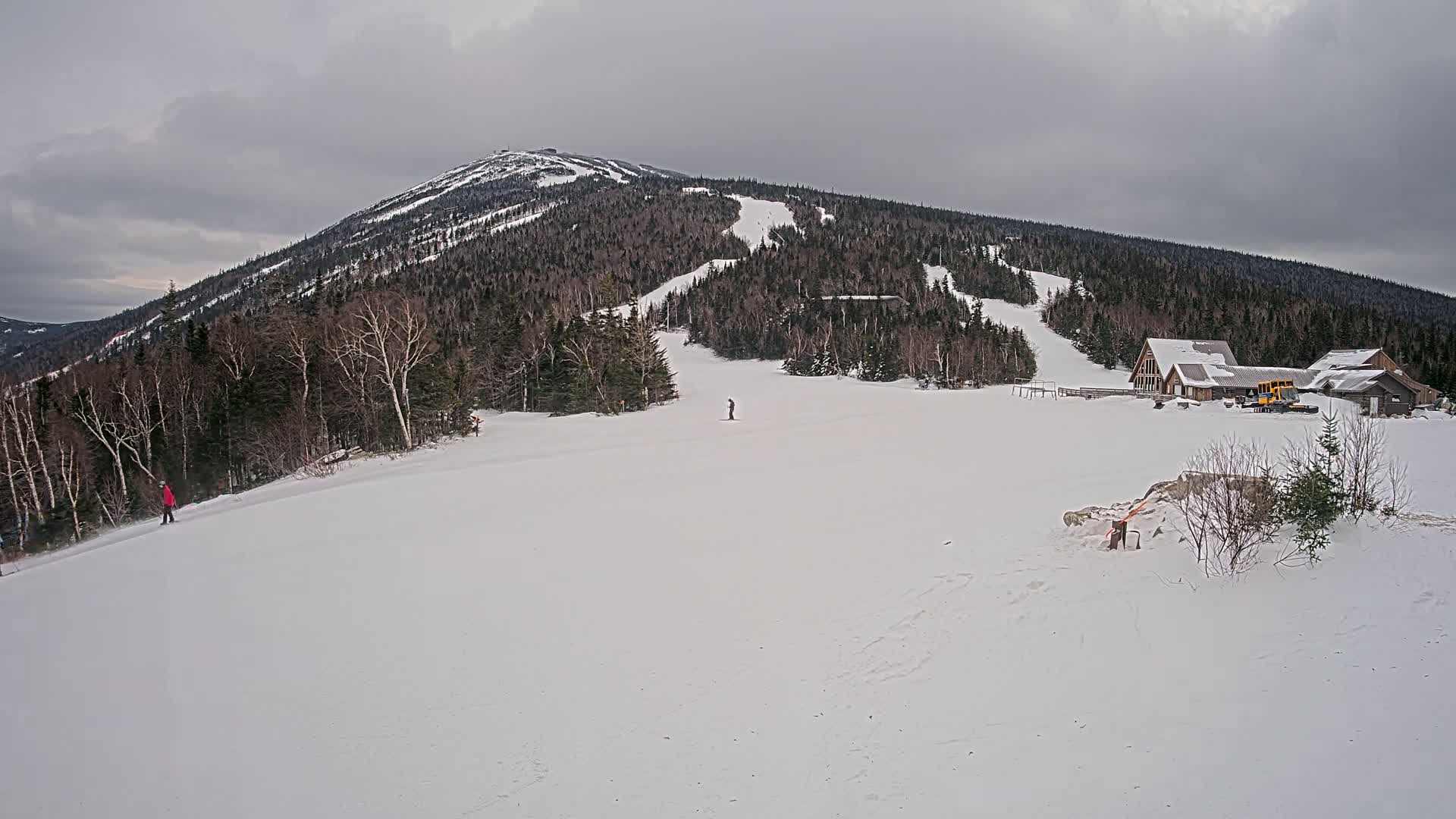A snow-covered mountain with ski slopes and a few skiers, under an overcast sky.