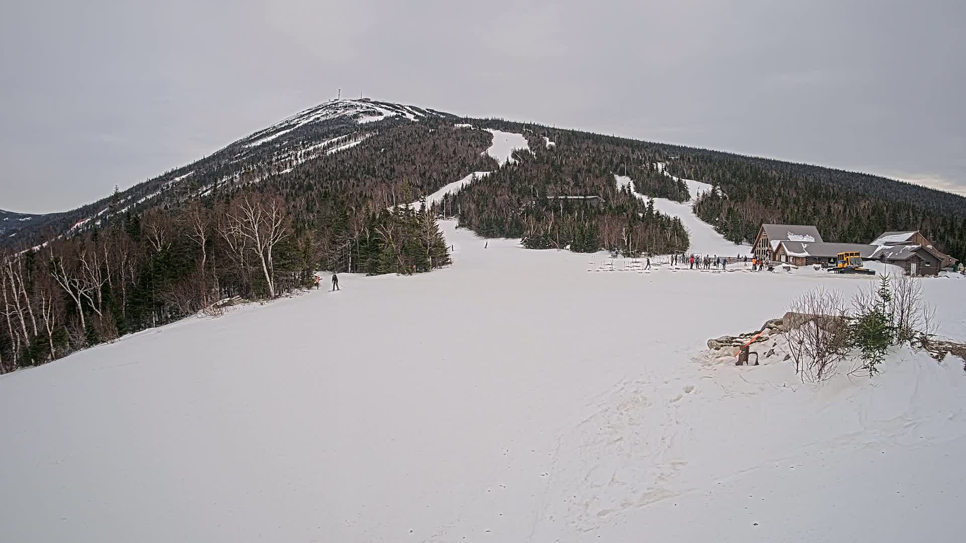 A snow-covered ski slope stretches from a base lodge toward a forested mountain under an overcast sky.