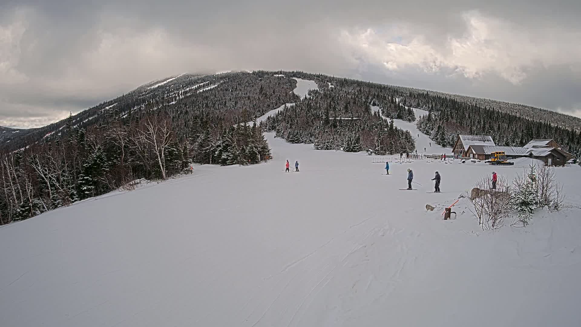 A snow-covered ski slope with several skiers and a snowcat near a cluster of buildings at the base of a wooded mountain under an overcast sky.