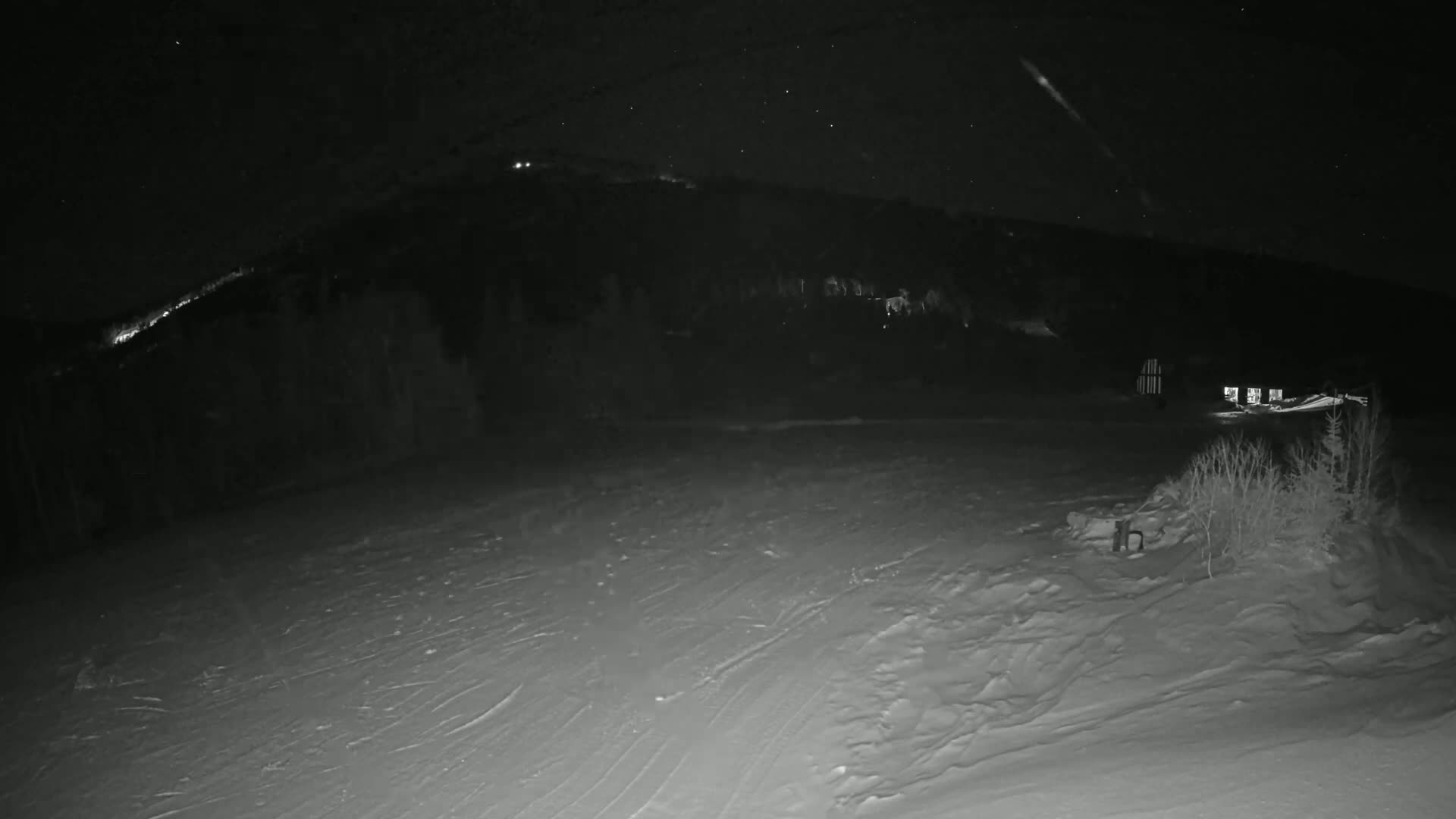 A snow-covered landscape at night, with a few buildings visible in the distance under a starry sky.