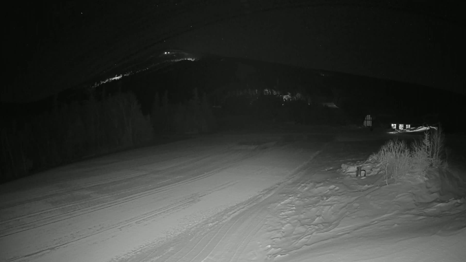 A snow-covered area at night, with a dark, snow-covered hillside and distant lights in the background.