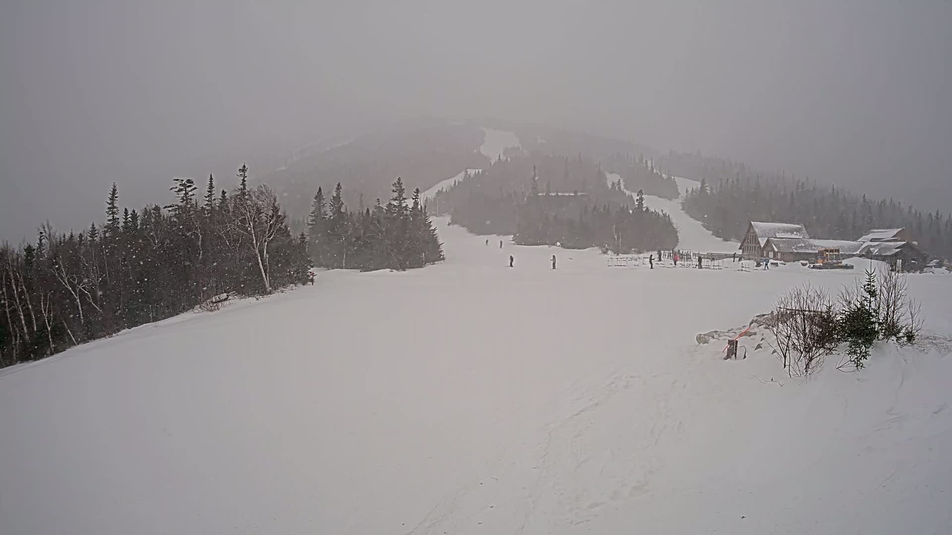 A snowy ski slope with a few skiers, a lodge, and evergreen trees is visible in heavy snowfall.