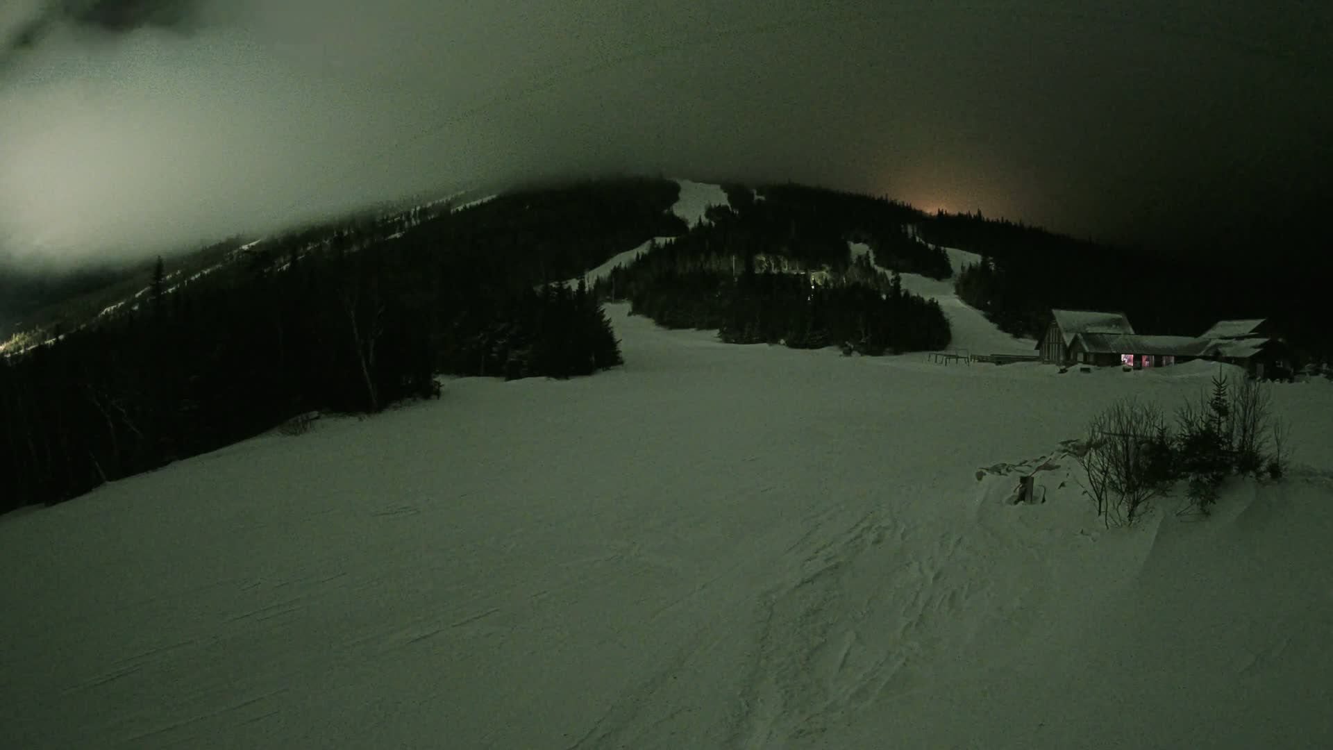 A snow-covered ski slope at night under a cloudy sky, with a building lit up in the distance.