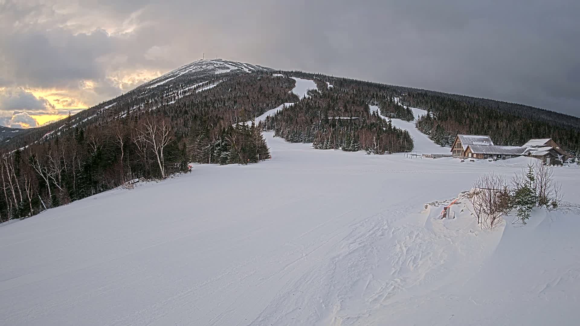 A snow-covered ski resort with a mountain in the background under a cloudy sky with some sunset hues.