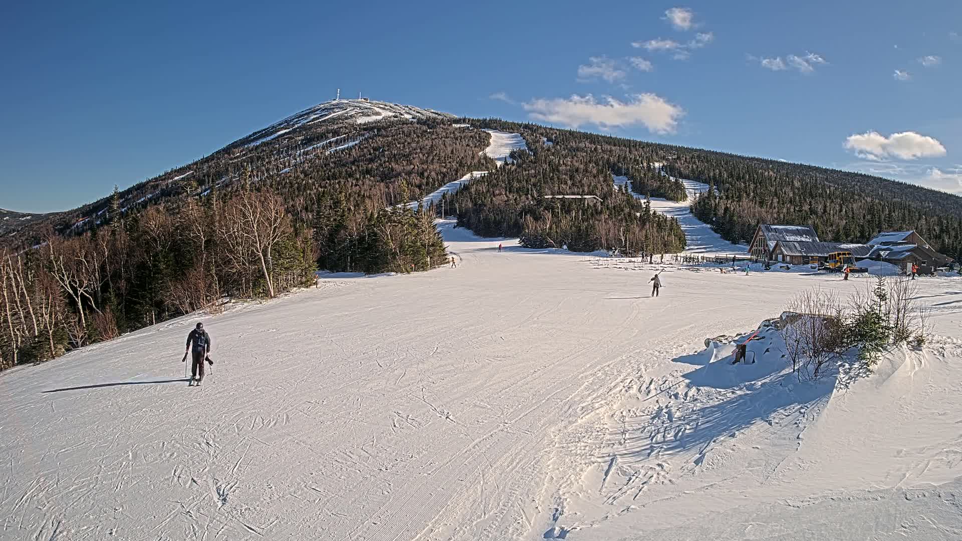 A snow-covered mountain slope with several skiers and snowboarders, a base lodge, and a clear blue sky.