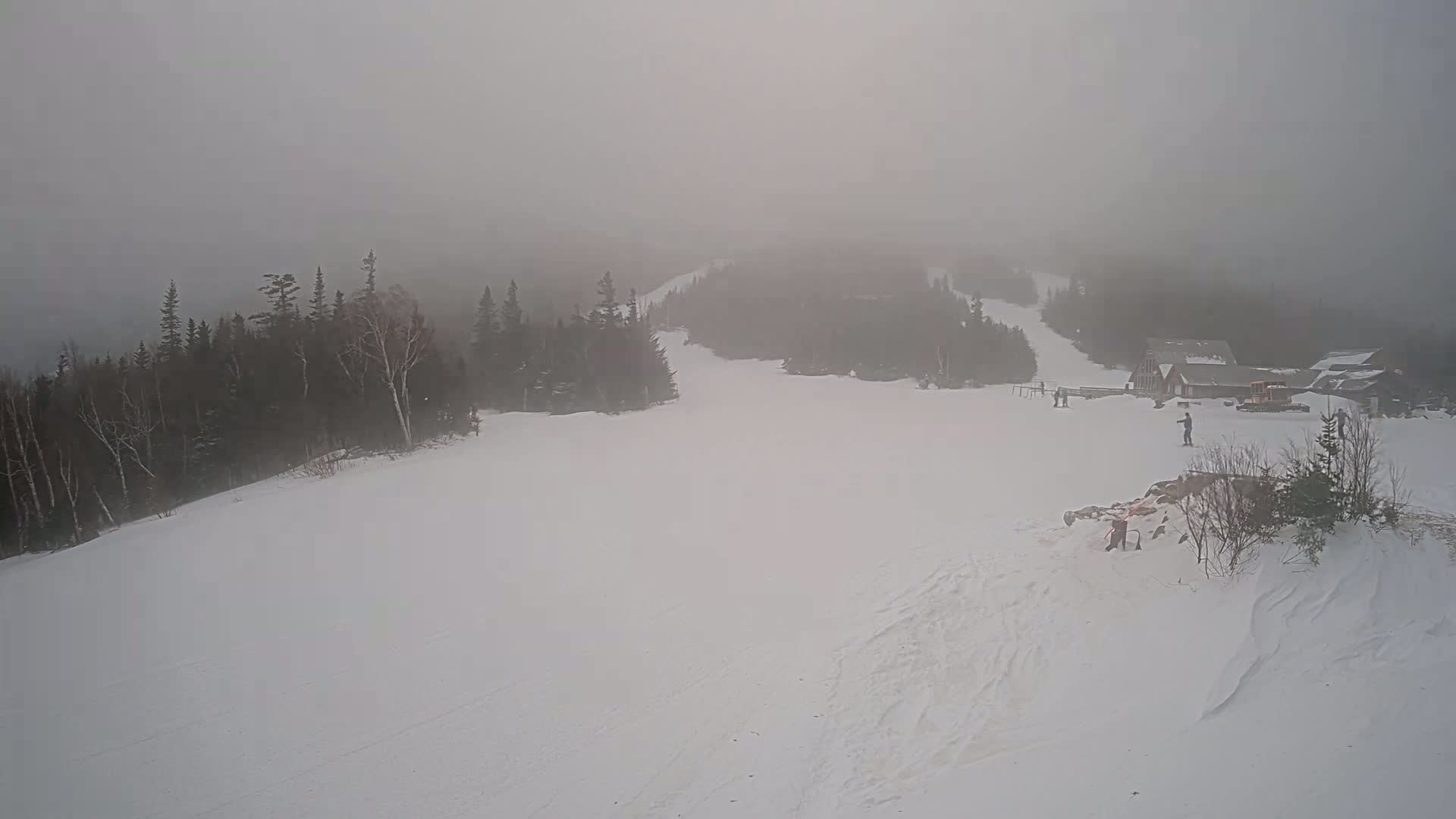 A snowy ski slope is shrouded in thick fog, with a few skiers visible near a cluster of buildings nestled amongst dark evergreen trees.