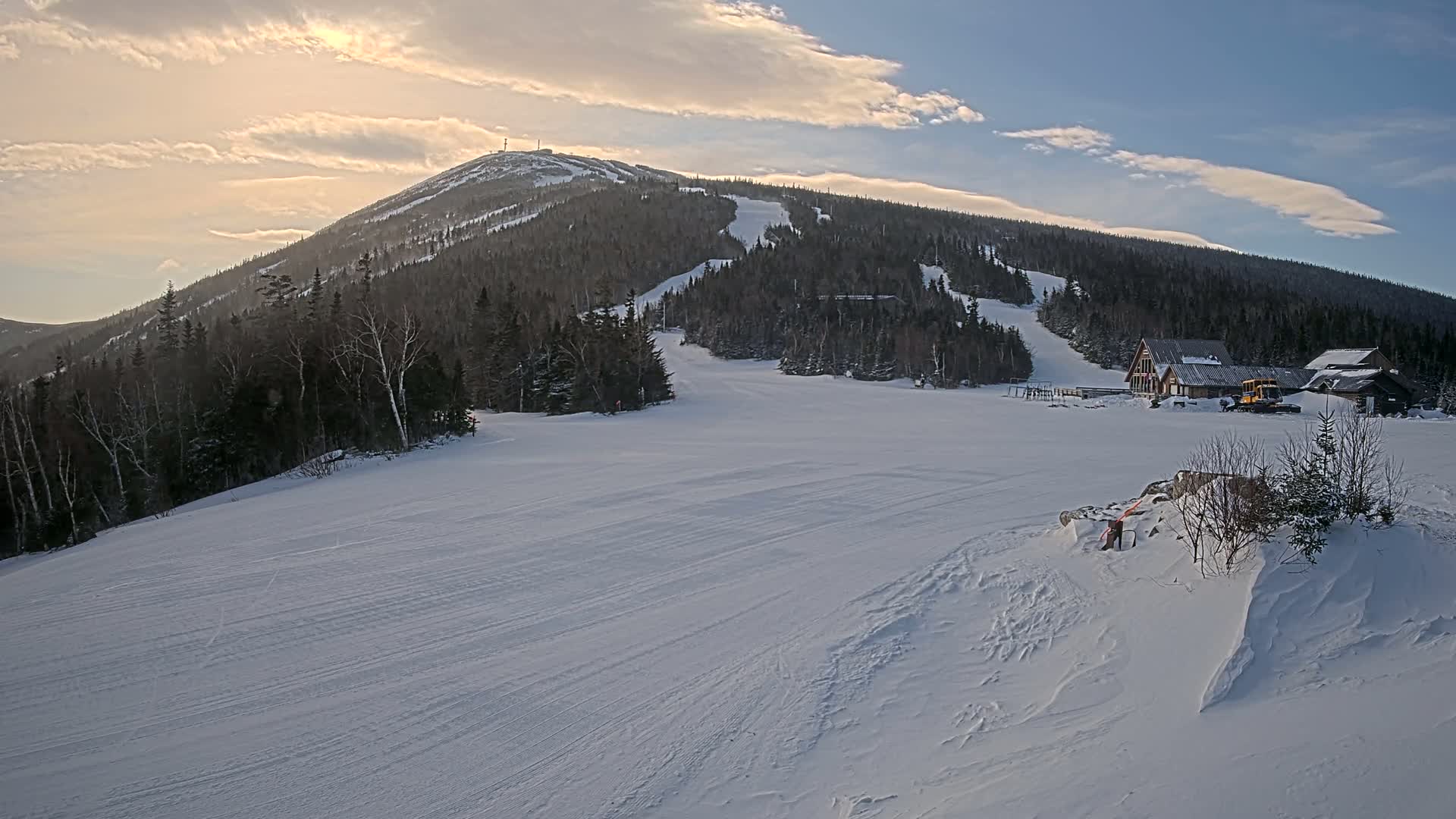 A snow-covered ski slope stretches out beneath a partly cloudy sky, with a mountain in the background featuring ski runs and a few buildings near the base.