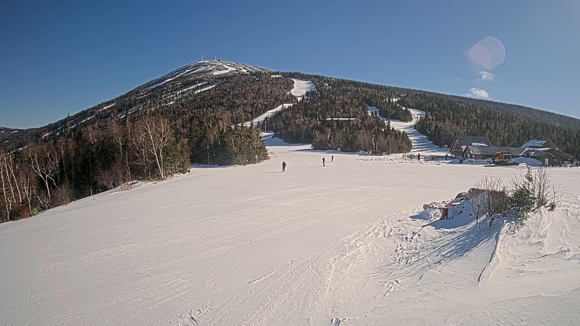 A snow-covered ski slope with several skiers, a ski lodge, and a mountain in the background under a clear blue sky.