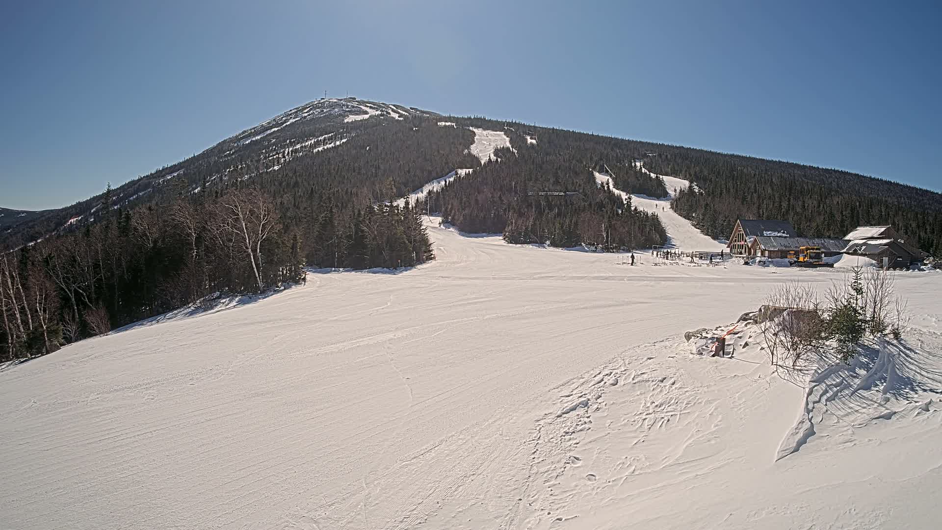 A snow-covered ski resort sits at the base of a large, forested mountain under a clear, sunny sky.