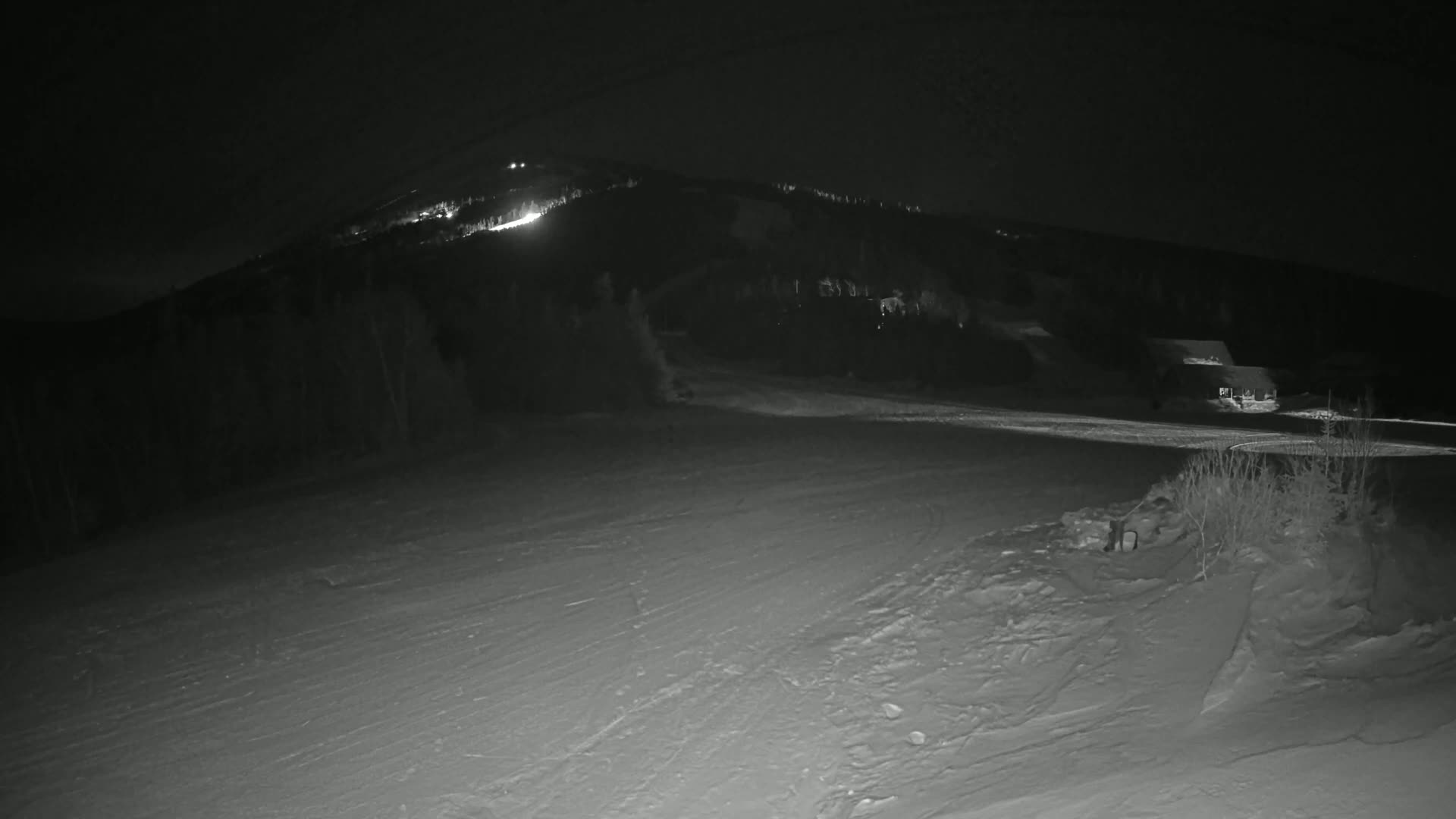 A snow-covered landscape at night, with a hill illuminated by lights in the distance.